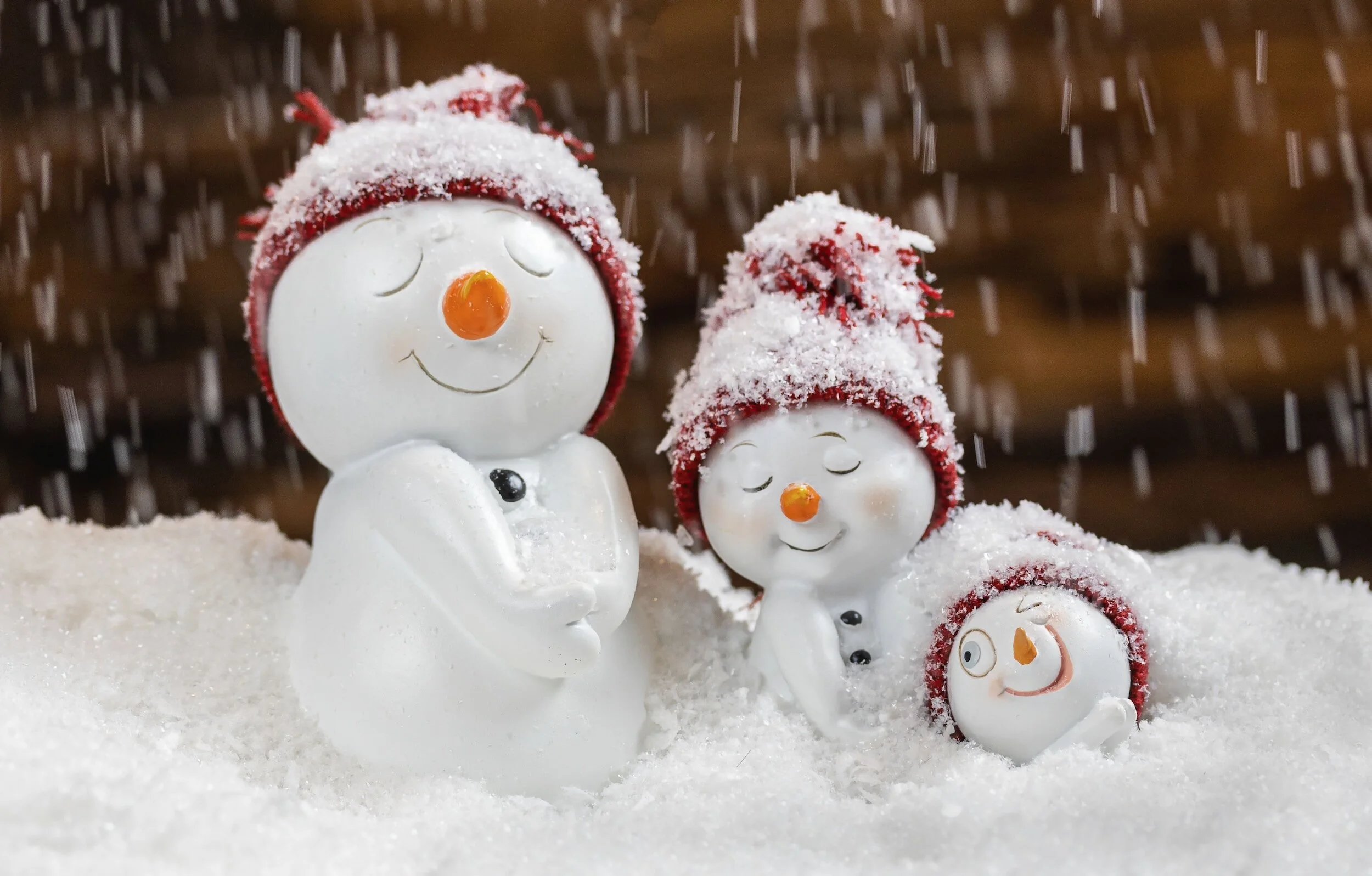 Large, medium, and small ceramic snowmen, wearing red knitted caps covered in snow, and nestled in more snow.