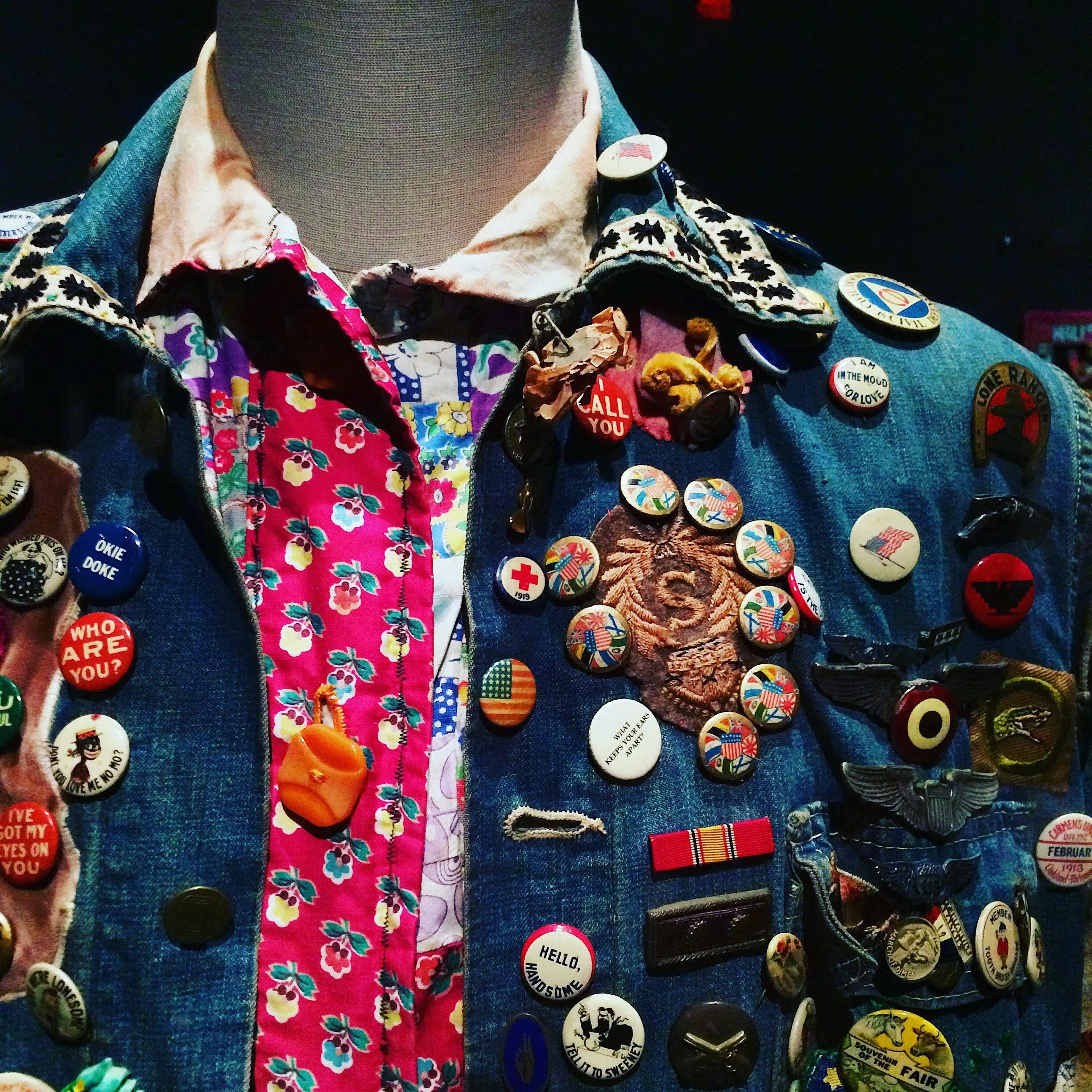 A jean jacket on a mannequin.  The jacket is covered in buttons and badges.