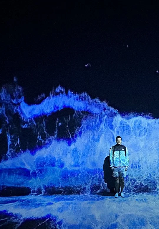 Person standing on a dark beach at night with blue illuminated ocean waves behind them.