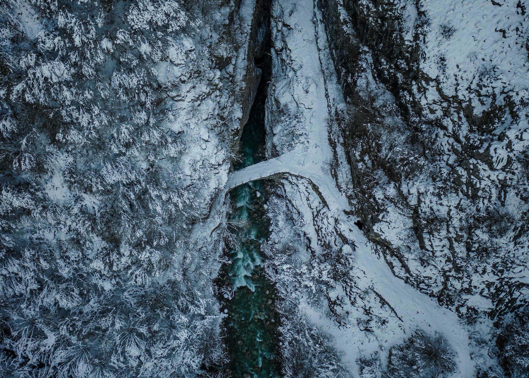 Aerial shot of a small bridge spanning a fast-flowing river in a steep canyon. The river is a vibrant turquoise, contrasting with the white snow and dark rocks of the gorge walls. Snow-covered trees line the edges of the canyon.