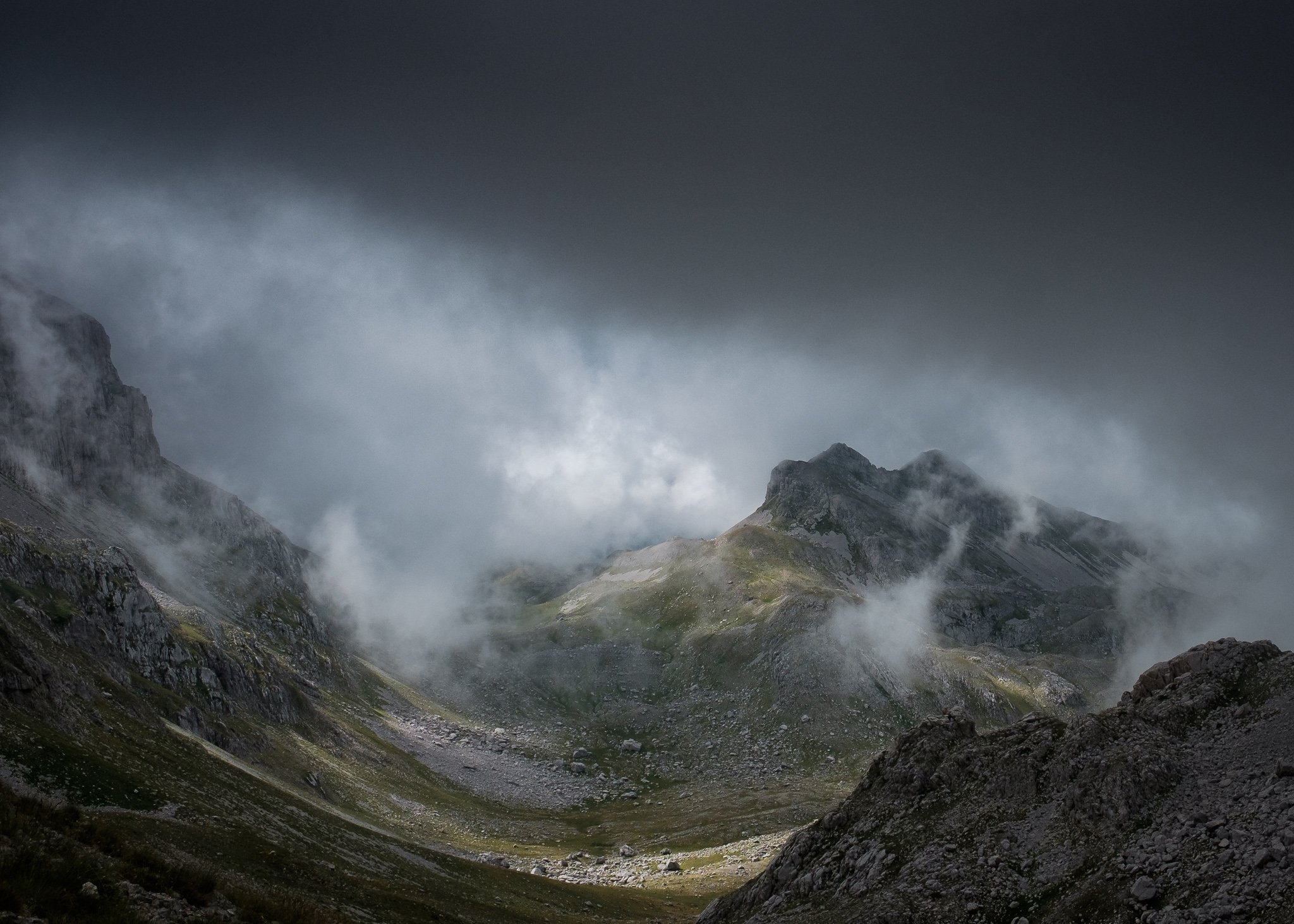 A dramatic mountain valley with mist rolling through steep slopes, patches of sunlight breaking through the heavy clouds, highlighting the terrain