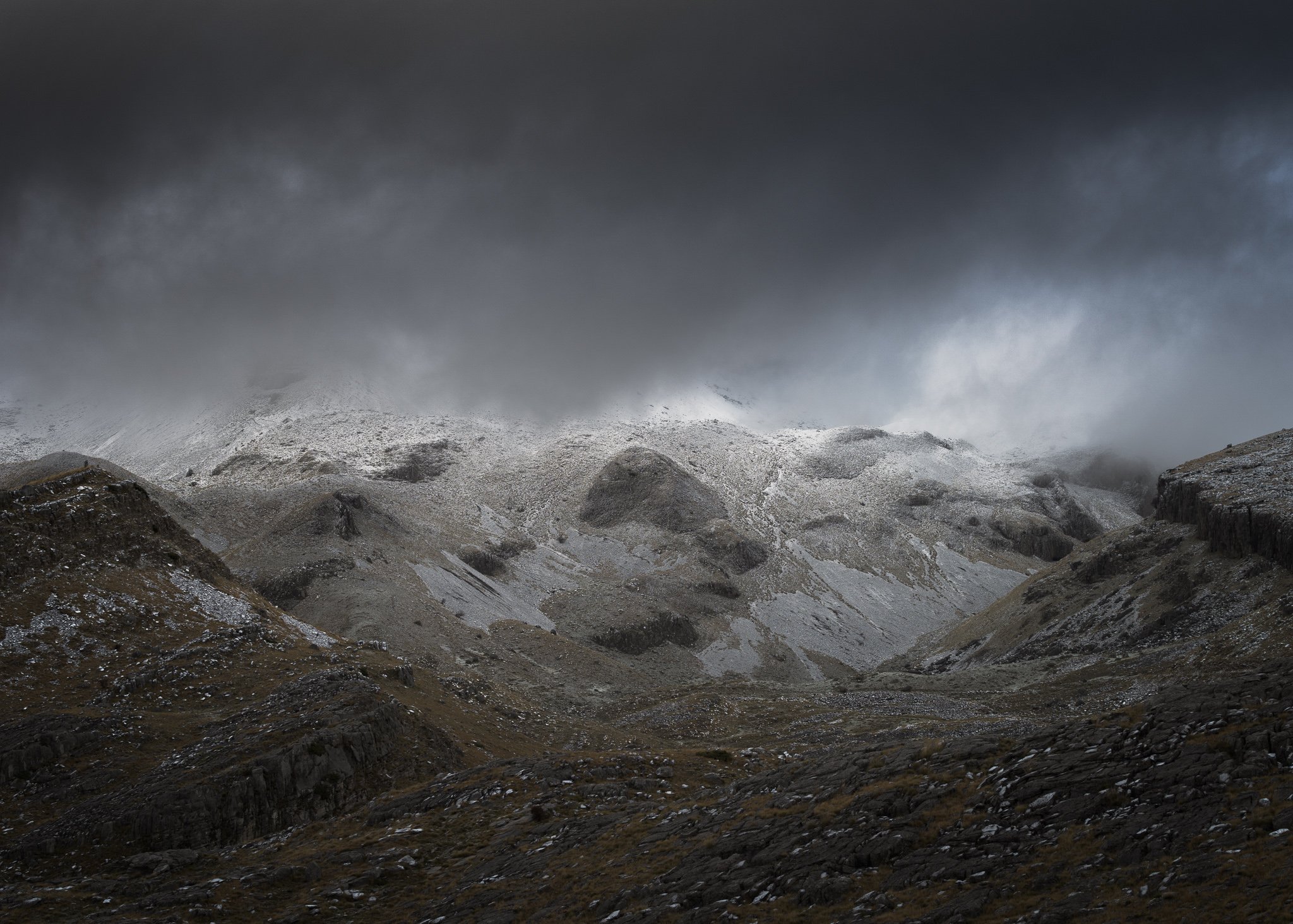 A landscape image of a mountain range with rocky outcrops and patches of snow, viewed under a dark and turbulent sky. The foreground is comprised of dark, rough terrain leading to the snow-covered peaks in the distance. The lighting is subdued, highl