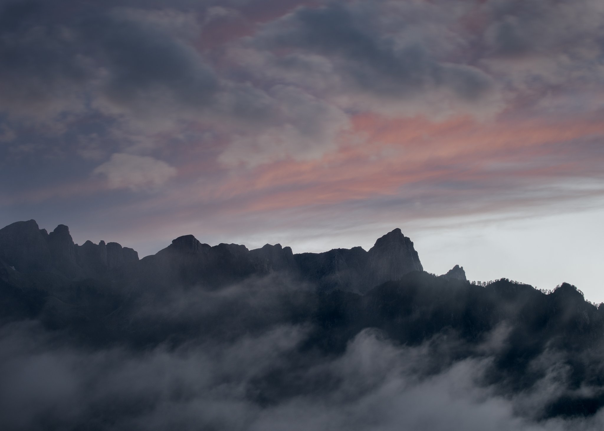 Silhouetted mountain peaks rise through low-hanging mist, set against a dramatic sky streaked with soft pink and blue hues at dusk.