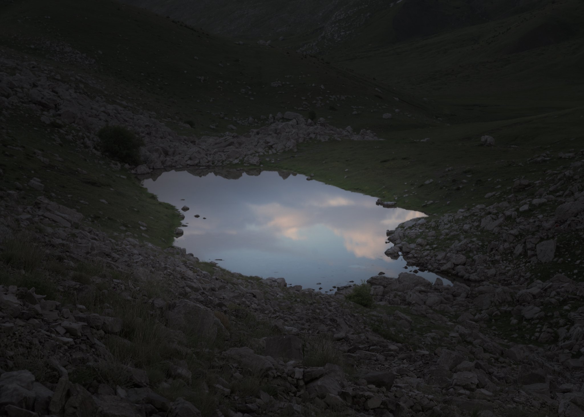 A small reflective alpine pond set among rocky terrain, mirroring soft pastel hues of the sky, contrasting with the rugged mountain slopes.