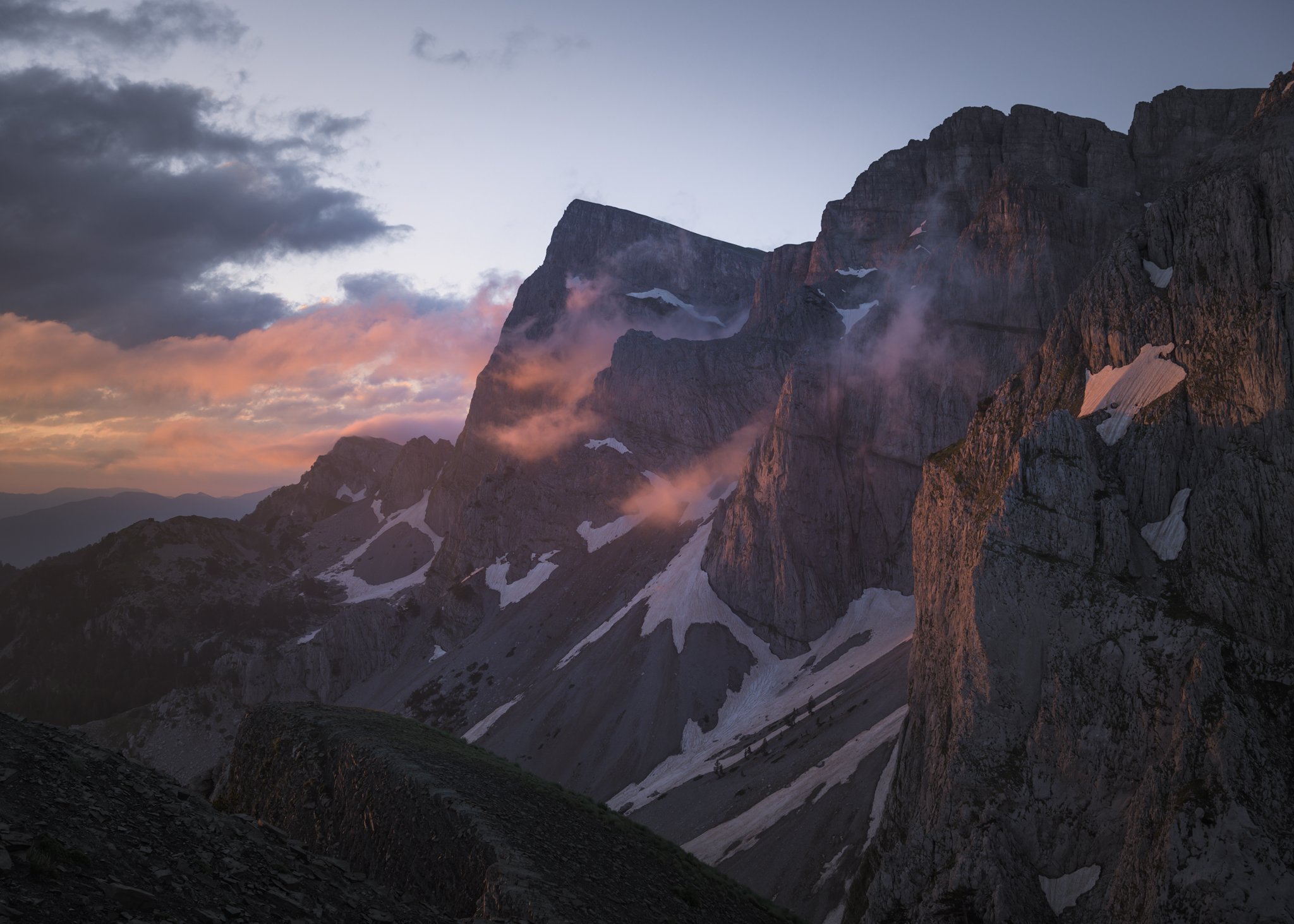 A towering mountain range bathed in warm sunset light, with vibrant pink-hued clouds drifting around the sheer rock faces and remnants of snow.