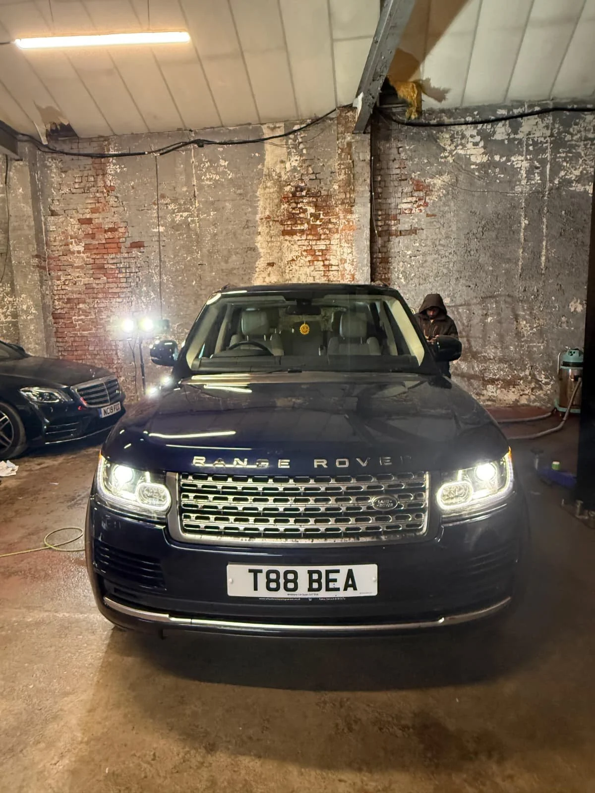 Front view of a black Range Rover with a UK license plate T88 BEA, parked indoors against a weathered brick and concrete wall, with another car visible to the left and a person in a dark hoodie on the right side.