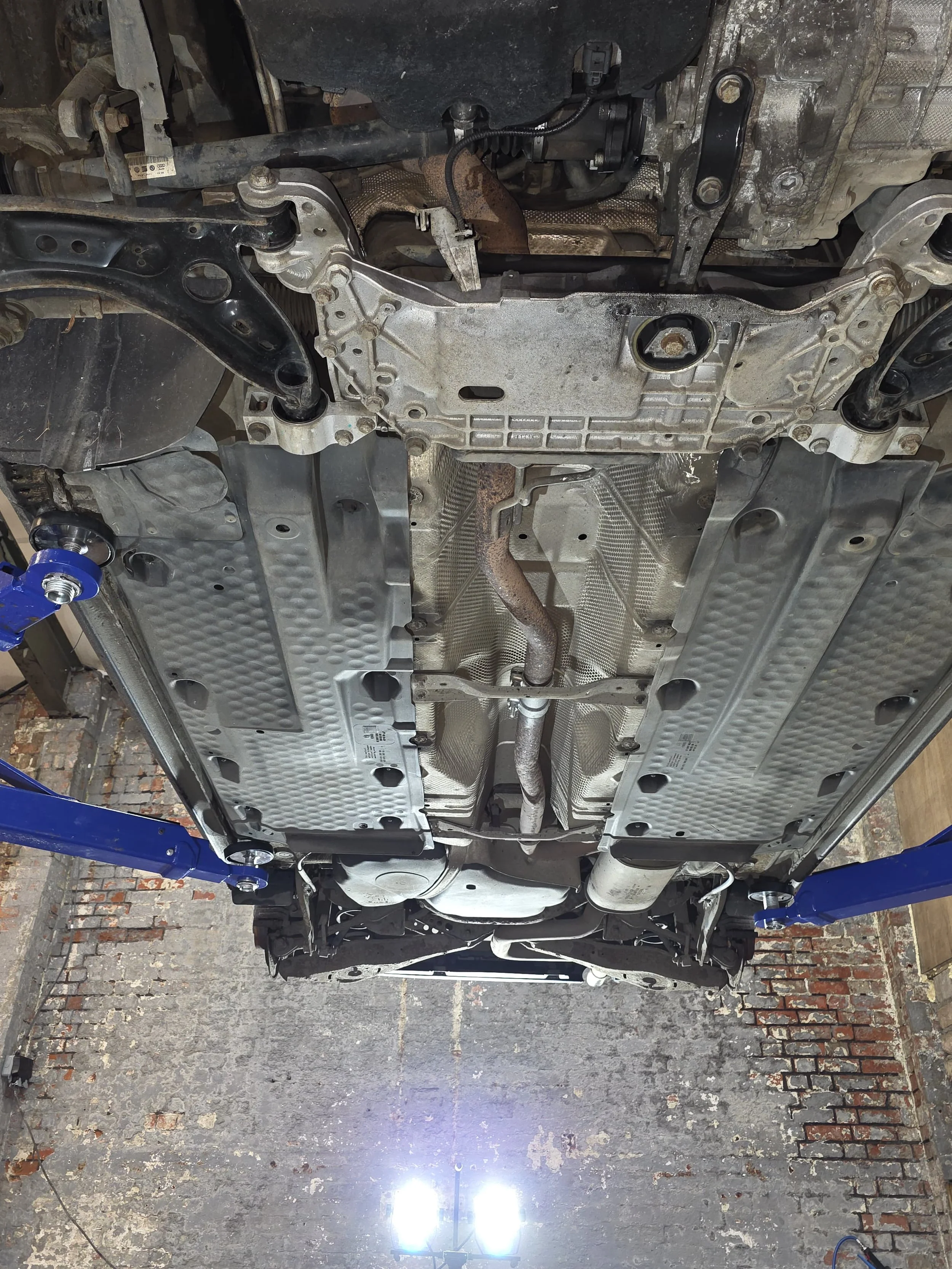 View of the underside of a car lifted on a hydraulic lift, showing the exhaust system, oil pan, and protective underbody shields.