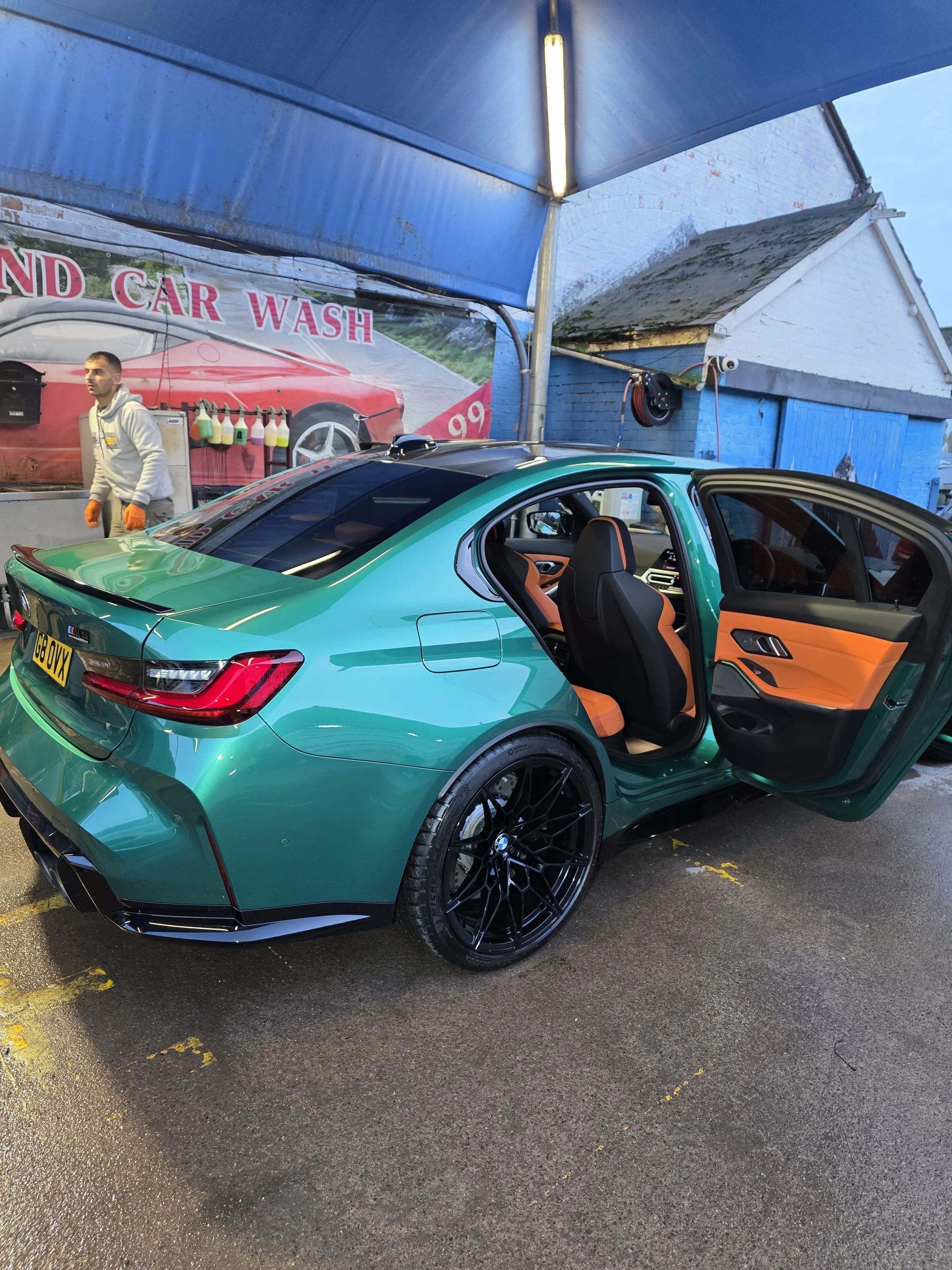 A teal-colored BMW sports car with black wheels parked at a car wash station, with the driver-side door open revealing a tan and black interior. A man in a gray hoodie and orange gloves stands nearby under a blue canopy, with a sign in the background