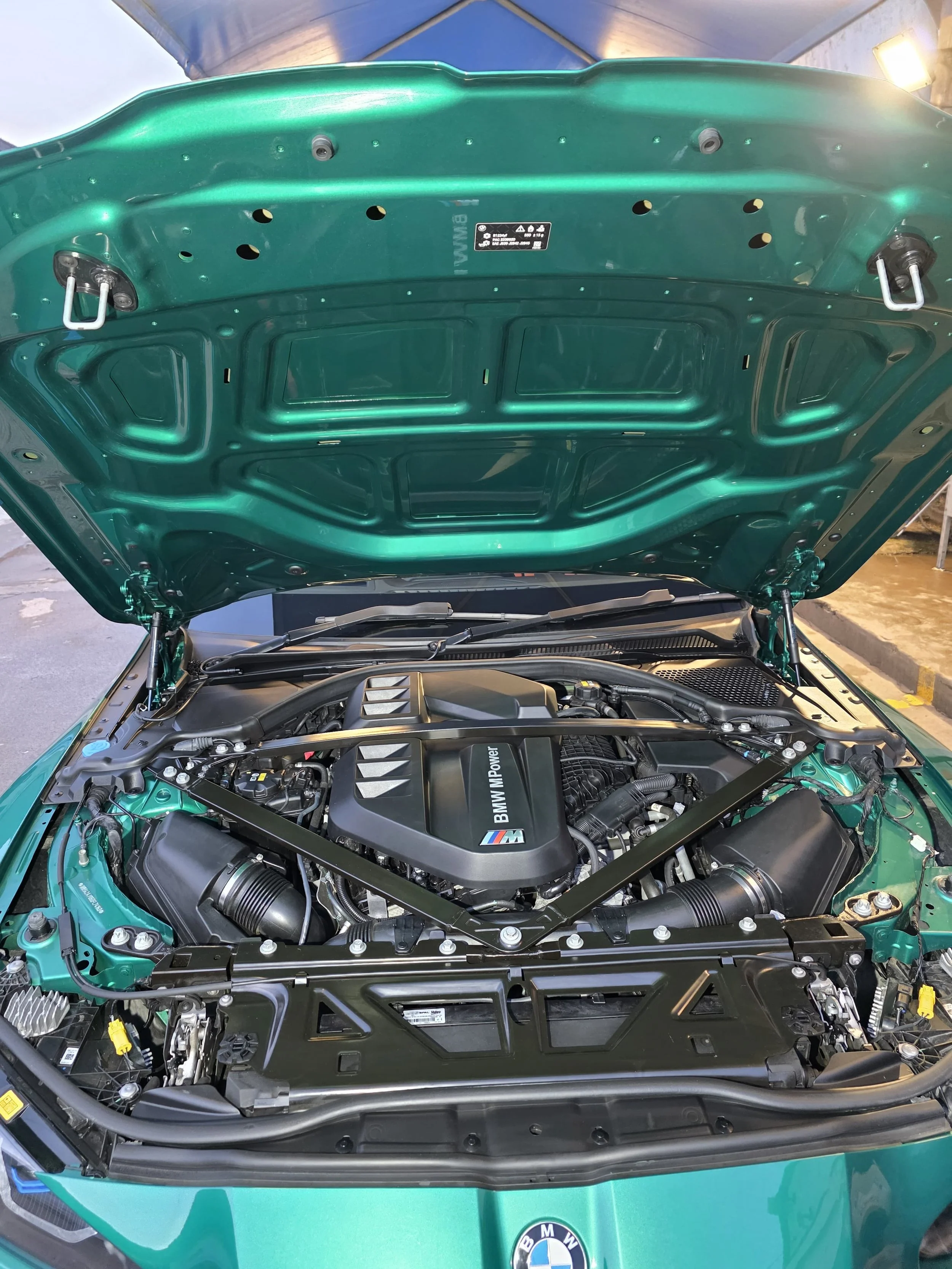 The image shows the open hood of a green BMW car, revealing its engine bay with a BMW M Power engine cover.