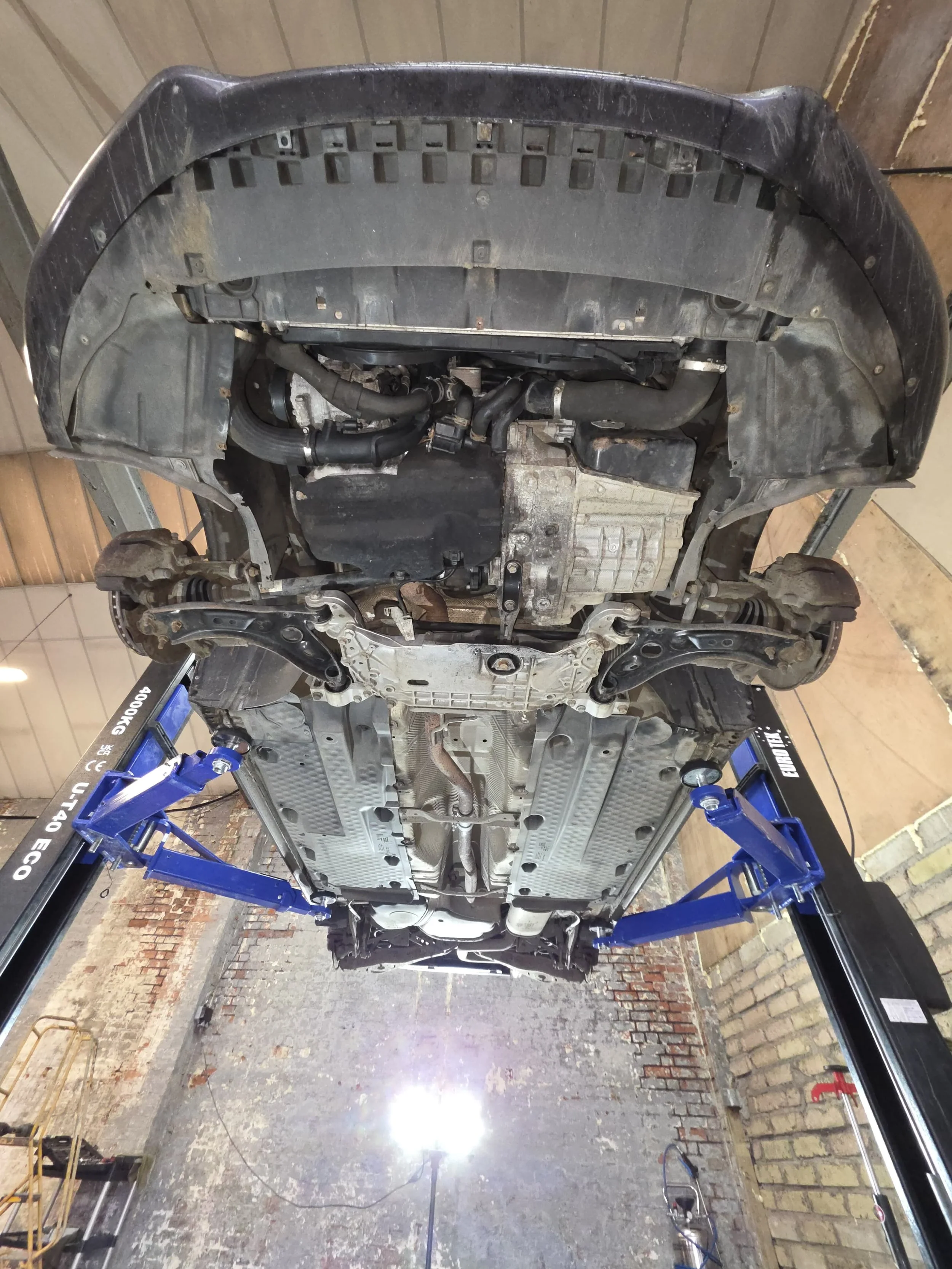 Underneath view of a car lifted on a hydraulic lift, showing the underside of the engine, suspension, and exhaust system.
