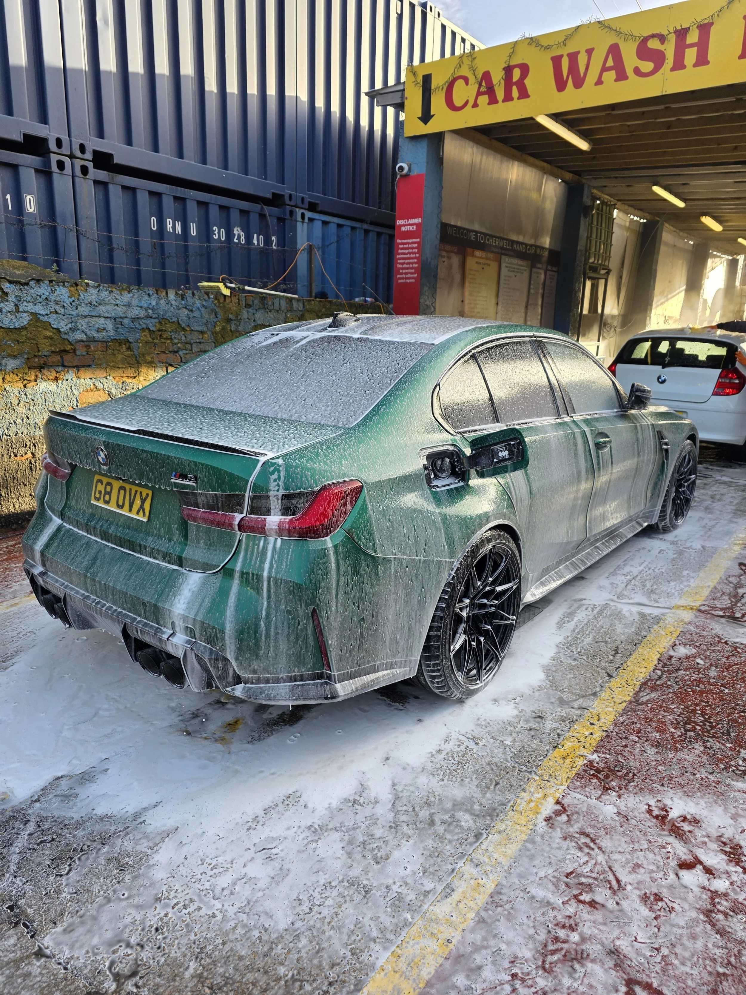 A green BMW M5 sports car covered in soap suds at a car wash bay, with other vehicles in the background.