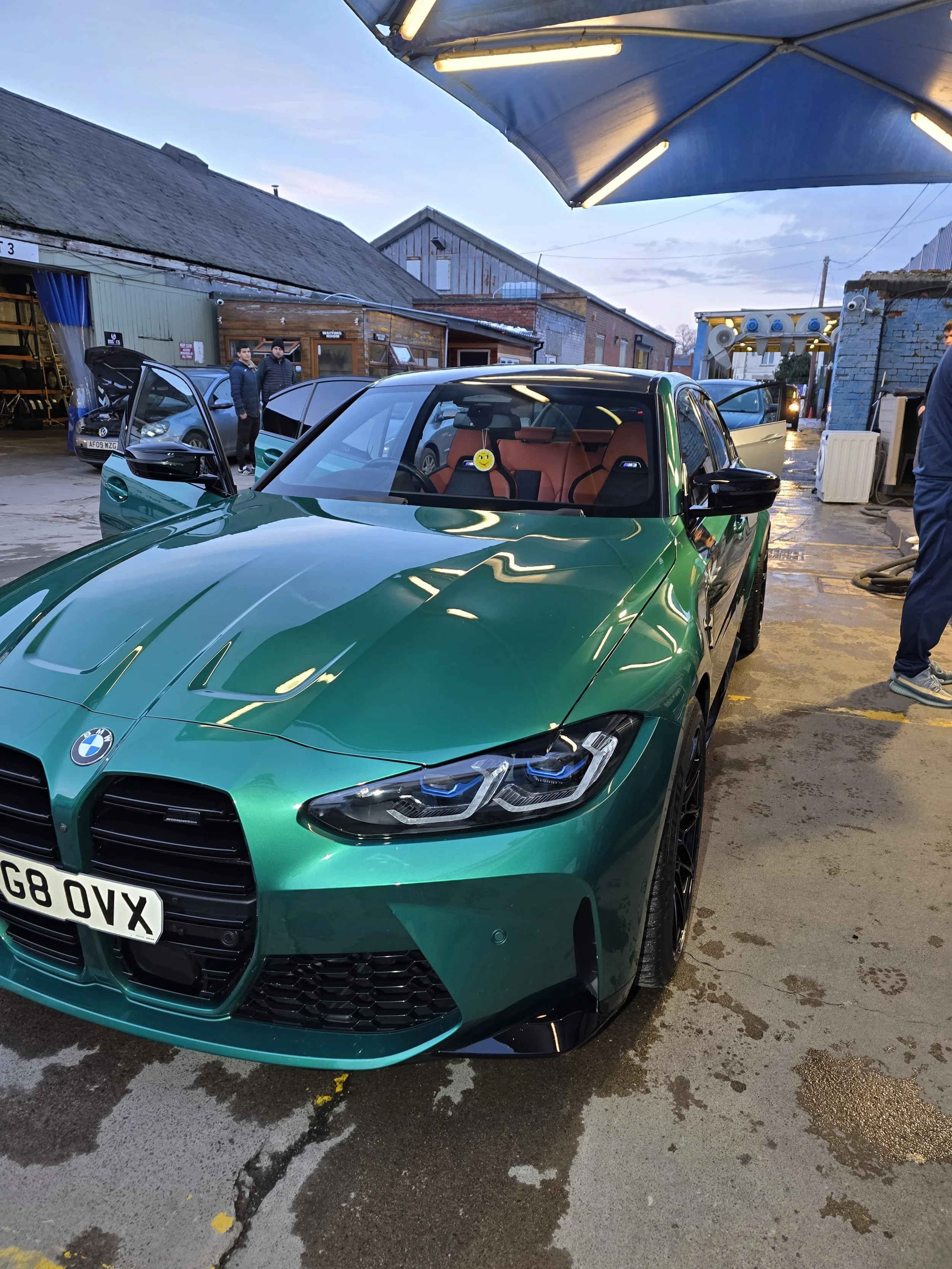 A green BMW car parked outdoors near a warehouse or garage with people in the background, and a large blue-and-white parasol overhead.