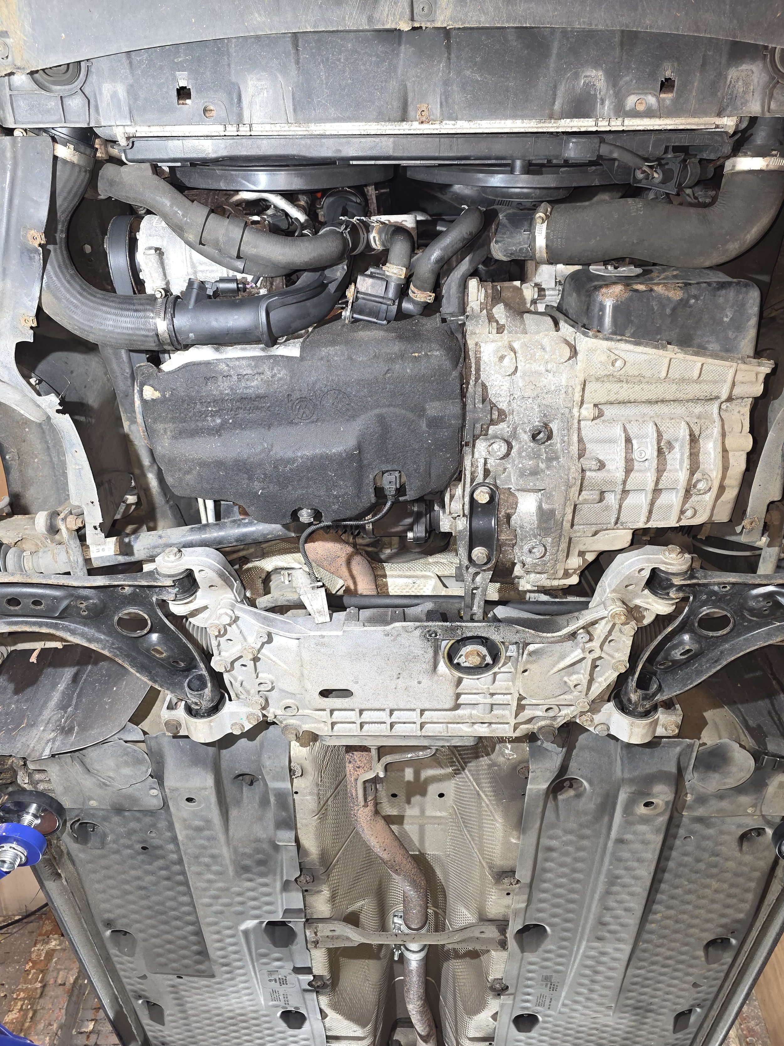 Underneath view of a car engine and front suspension, showing various engine components, hoses, and metal parts.
