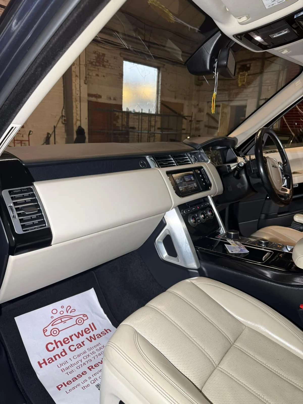 The interior of a vehicle showing the dashboard, steering wheel, and front seat. A sign on the floor reads 'Cherwell Hand Car Wash' with an address and phone number.