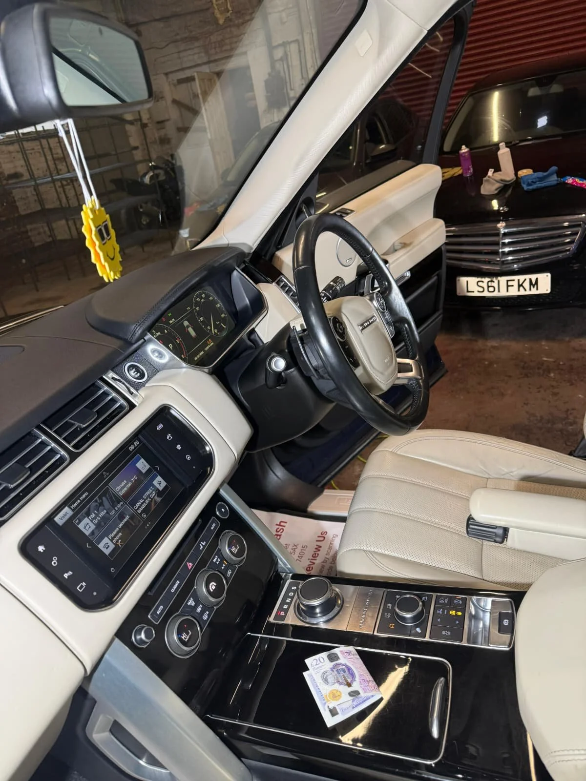 The interior of a luxury vehicle with beige leather seats, a touchscreen display, and control dials, with a UK twenty-pound banknote on the center console. The dashboard shows various controls and the steering wheel is visible.