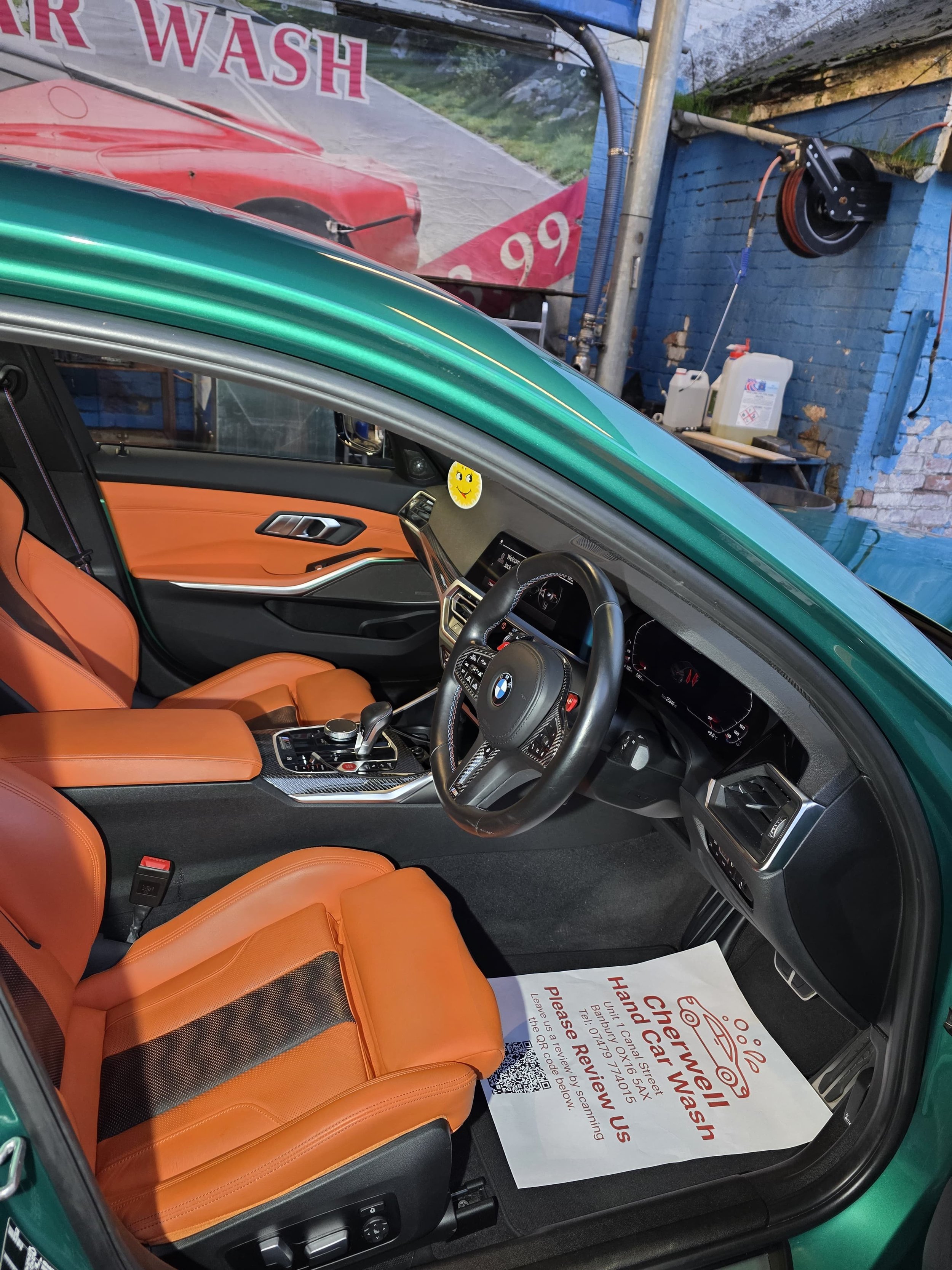 Interior of a green BMW car with tan leather seats, showing the dashboard, steering wheel, gear shift, and a moving control knob. The car is parked at a car wash, with a sign on the floor mat that reads 'Thank You, Hand Wash, Please Review Us' with a