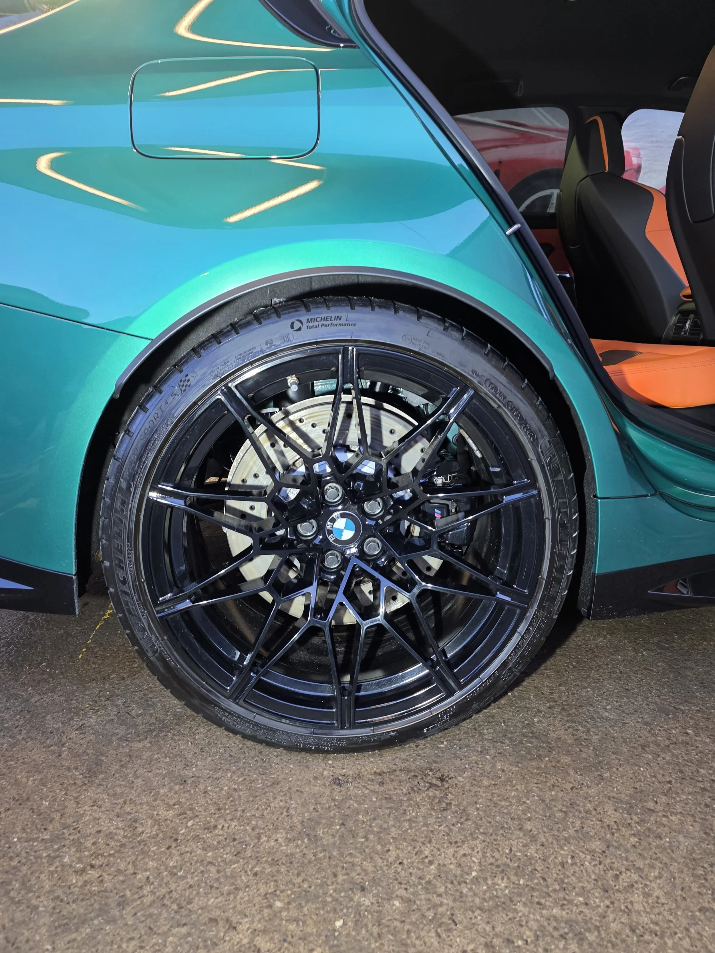 Close-up of a car's rear wheel with black, multi-spoke rim and Michelin tire, parked on a textured surface, with part of the turquoise body and an open door showing orange interior detail.