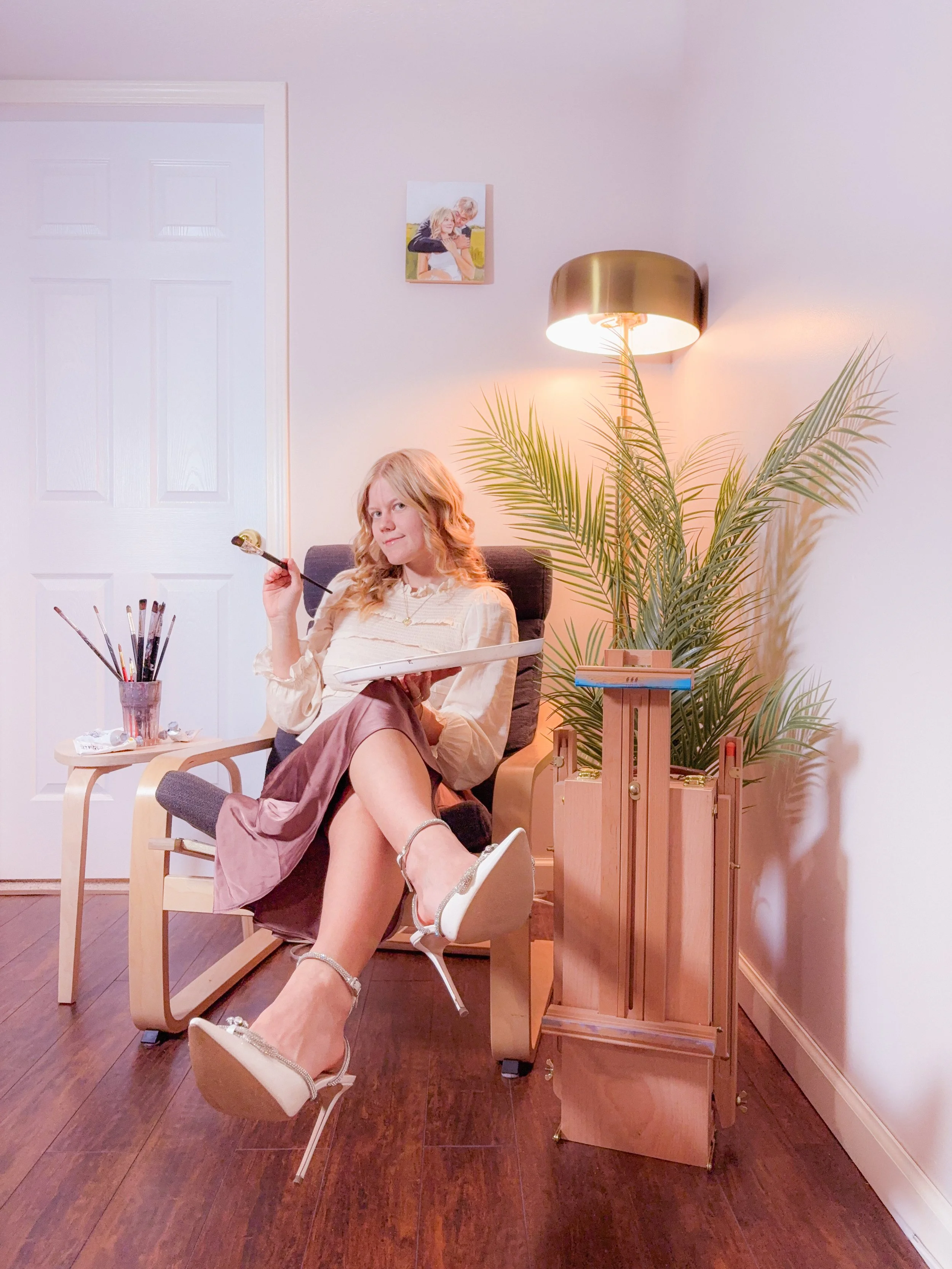 A woman sitting in a black and light wood lounge chair, holding a paintbrush and a palette, with a small table with paint supplies next to her, surrounded by a large plant and a wall lamp in a cozy indoor space.