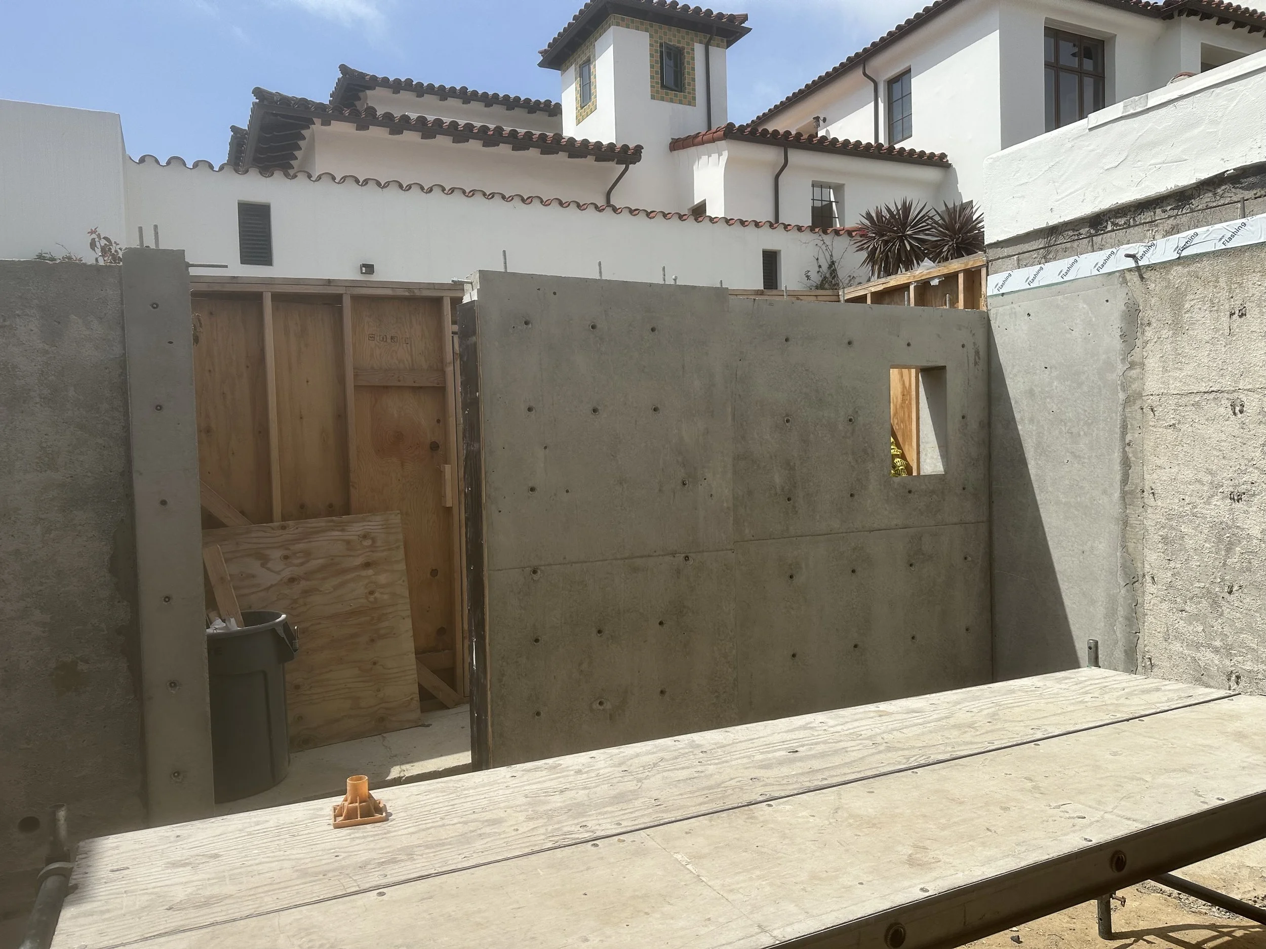  Exterior construction shot: a newly poured concrete wall section forms a small enclosure with a plywood-framed door opening. In the background, the existing Spanish-tile-roofed museum building is visible over the wall. 