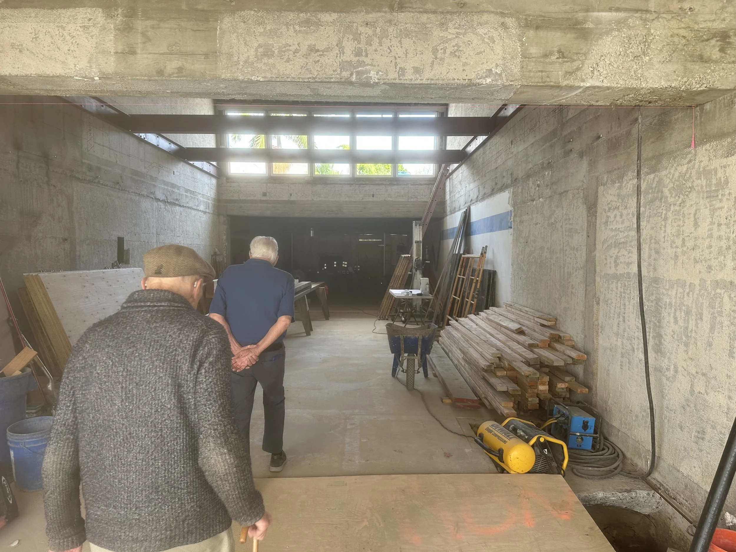  Interior of a poured-concrete structure, mid-construction, with clerestory windows letting in daylight near the ceiling. Two men walk toward the back of the space: Museum Staff (wearing a flat cap, using a cane) and Art Bosse, the museum’s owner (in