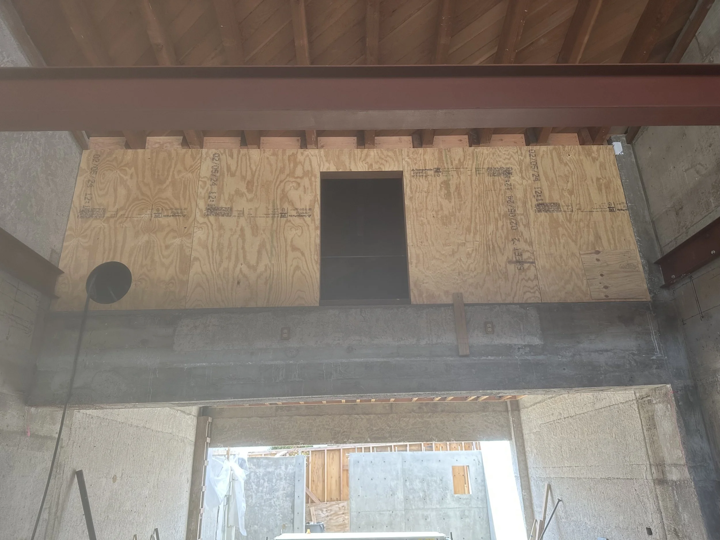  Looking up at the framing for the “boxcar” — the HVAC enclosure: plywood decking sits between wood roof trusses, with a steel beam running horizontally. Below, a concrete beam spans an open doorway leading outside. 