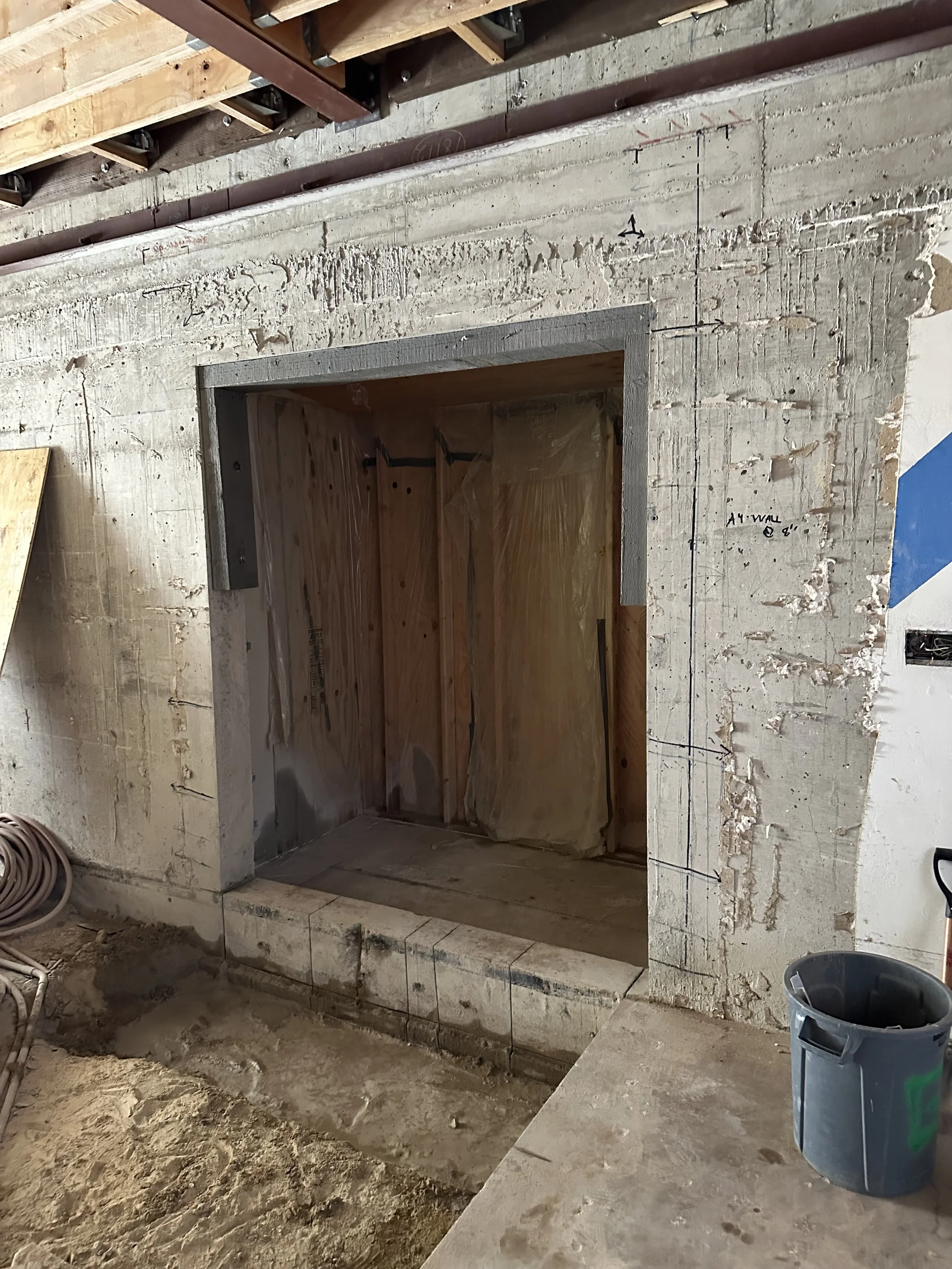  Construction progress shot looking into a framed wall opening/window cavity wrapped in plastic sheeting, poured-concrete walls surrounding it, with a gray duct/vent pipe visible nearby. 