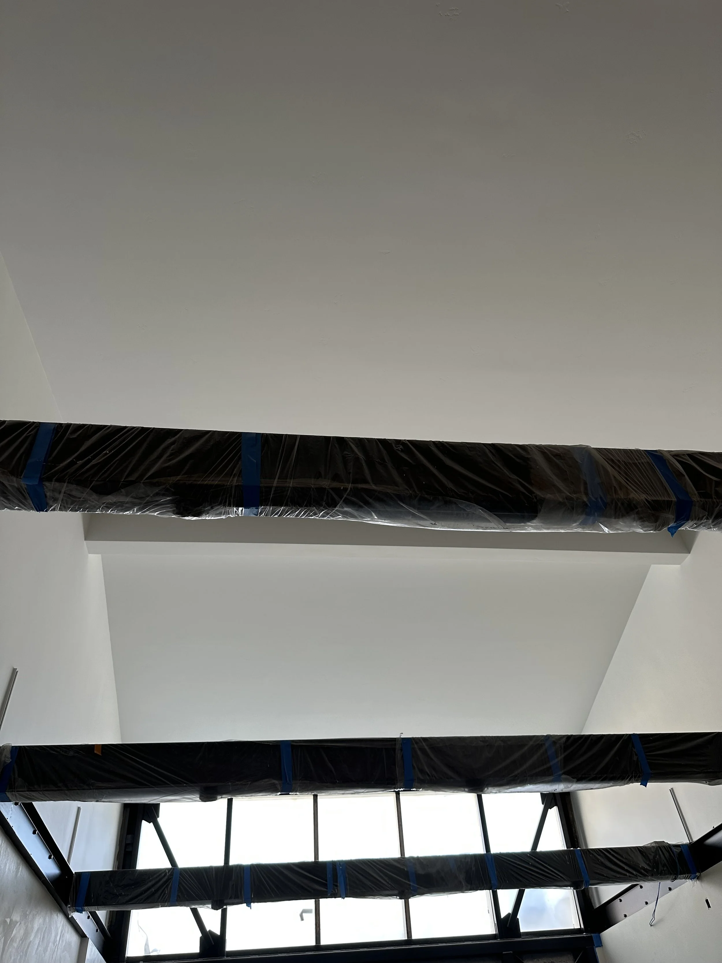  Two support columns wrapped in black plastic sheeting inside a bright, white-drywalled finished room, tall glass windows. 