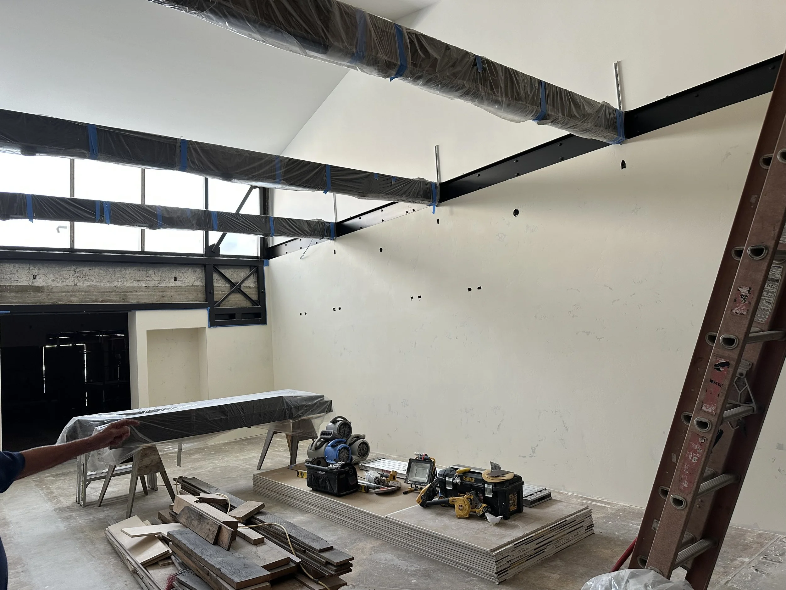  Large open room interior with white finished walls and exposed steel beams wrapped in plastic sheeting, tall windows. 