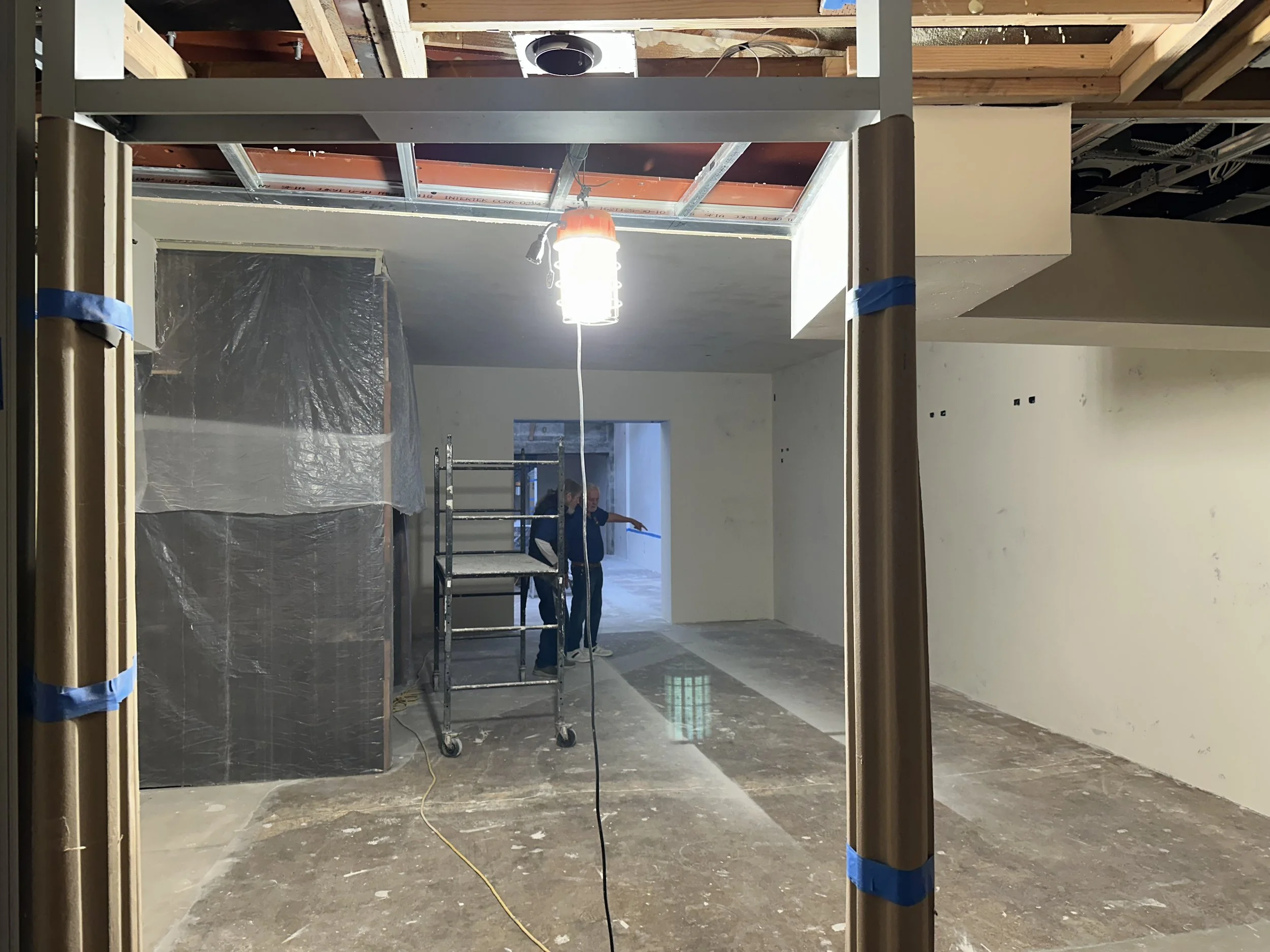  Interior room nearing completion: Museum Staff talk near a rolling scaffold, with a plywood ceiling patch overhead. 