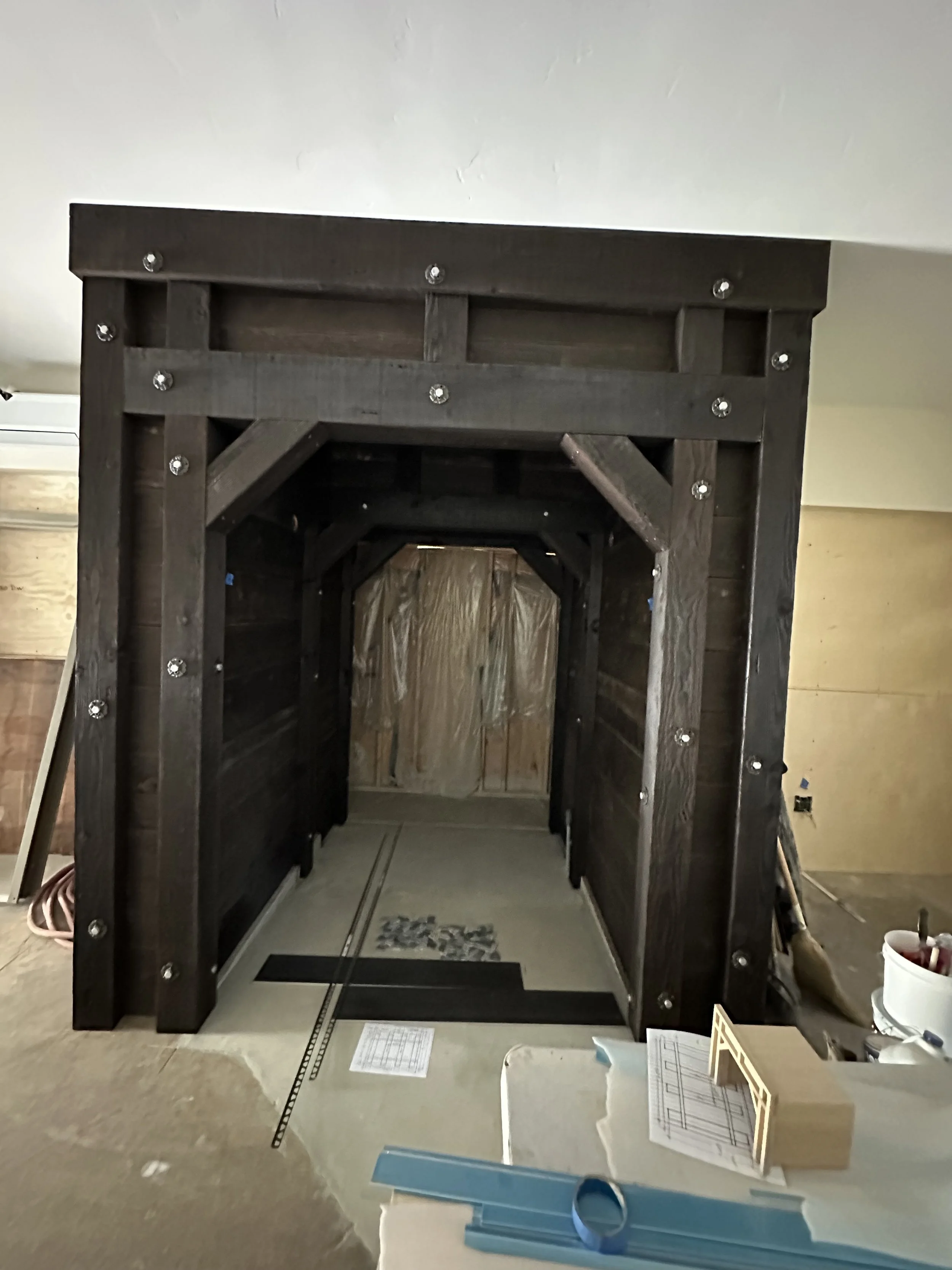  Construction progress shot: a heavy dark-stained timber-framed opening/portal leading into a room with materials stacked against the far wall. A framing detail sheet is posted on the wall nearby. 