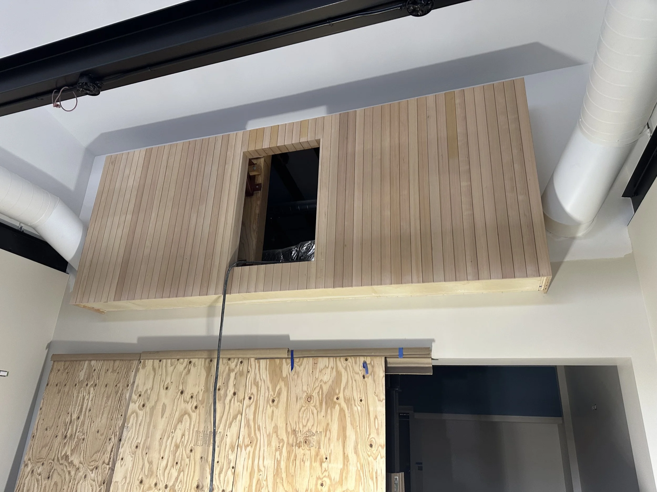  The “boxcar” HVAC enclosure, further along than the March 2025 framing shot: now clad in vertical wood slats, mounted high on the wall with a small square access opening. 