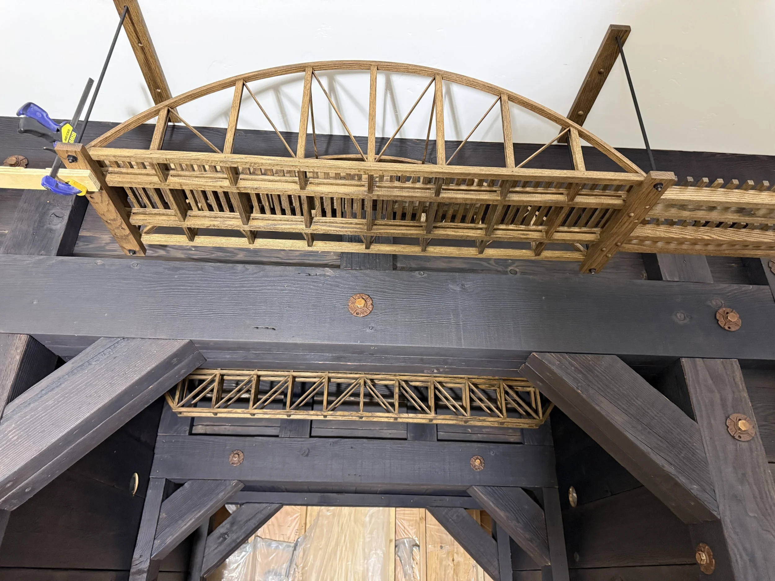  Close-up of a wood-truss arch bridge section of the overhead track, clamped in place during installation, with a smaller truss bridge segment visible below on a lower track run. 