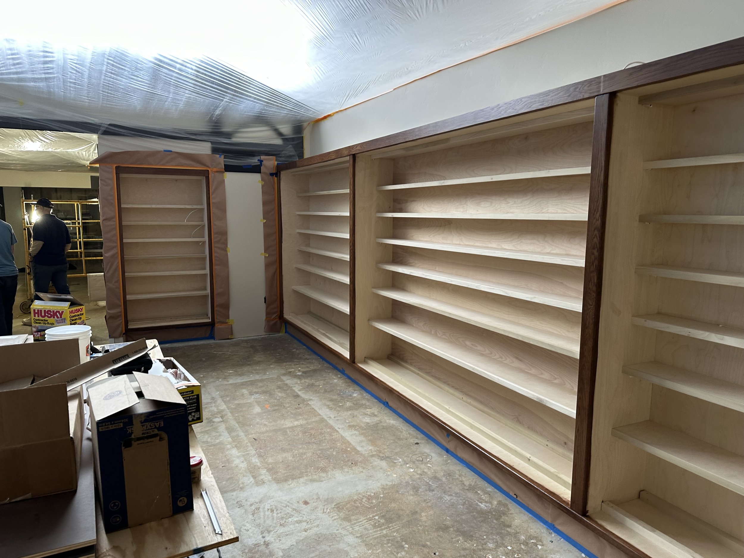  Wide shot of a room lined with the new built-in display shelving on multiple walls; Museum Staff confer near scaffolding at the back of the room. 