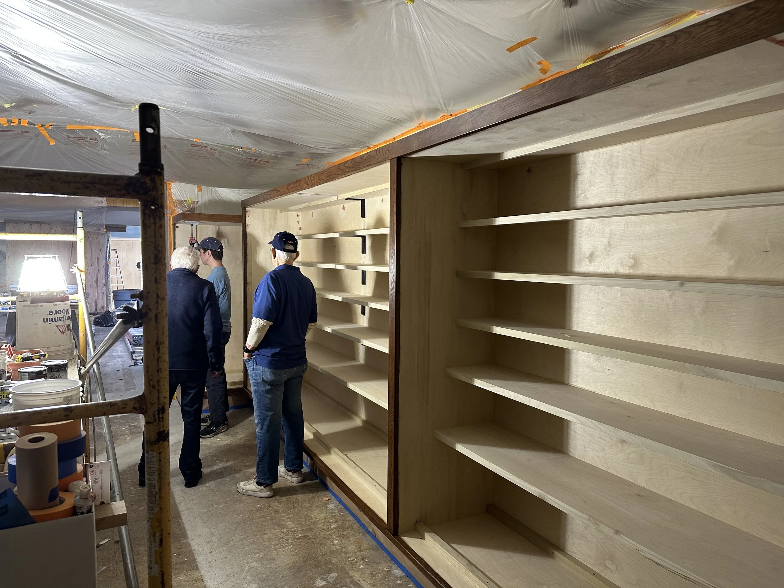  Museum Staff stand looking up at and inspecting a run of the new built-in shelving, mid-installation. 