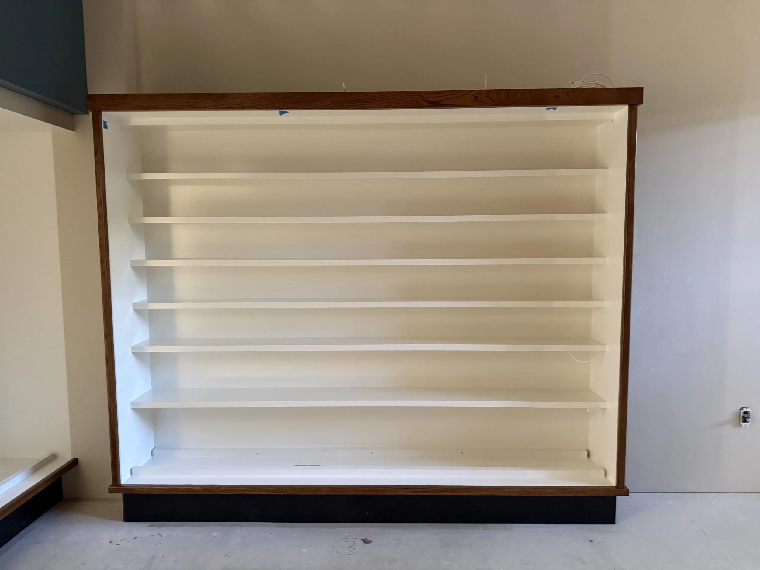  Wide shot of a finished built-in shelving unit — white-painted shelves in a dark wood-trimmed frame, empty and ready for display pieces. 