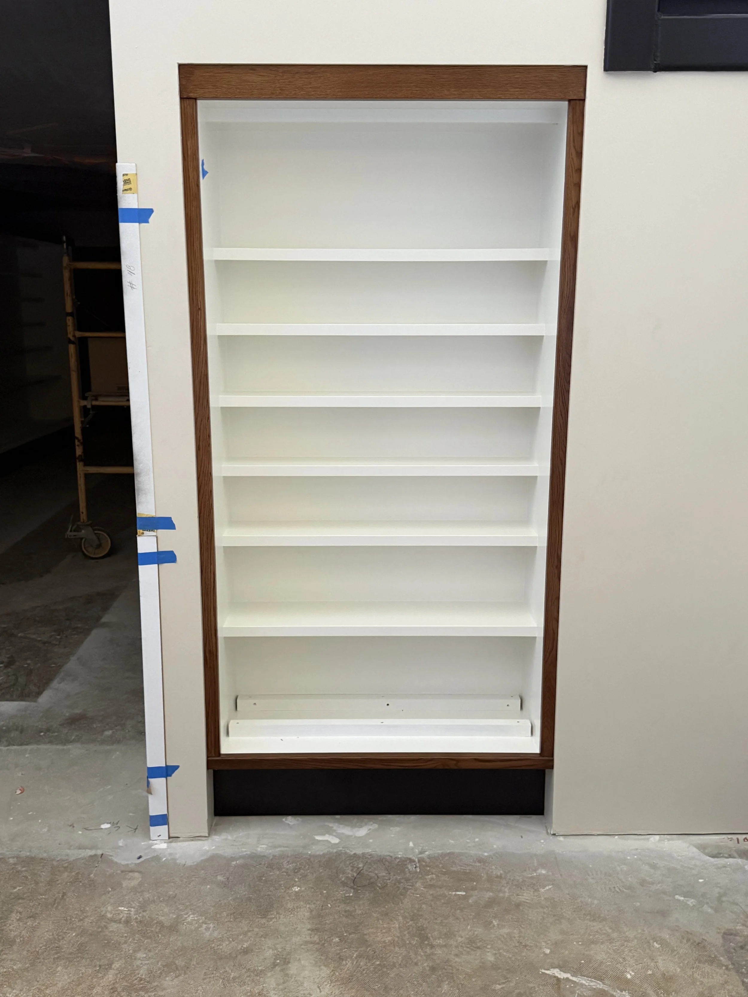  Vertical shelving unit with narrow divided compartments, wood-trimmed frame, mounted in a wall niche. 