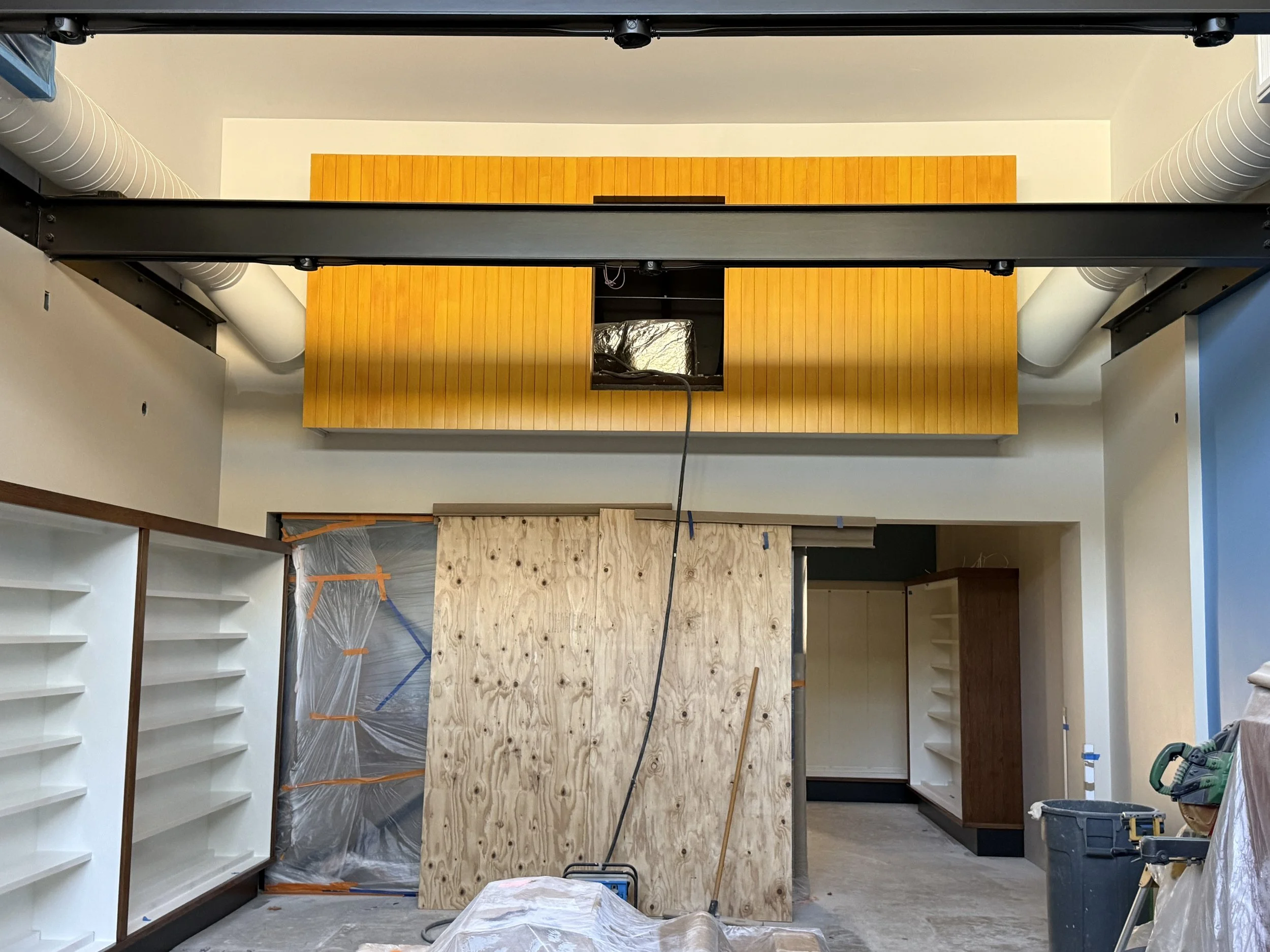  Wide interior shot: the boxcar HVAC enclosure, now finished in mustard-yellow wood cladding with an open access panel, hangs above a room under active work — plywood panel, shelving bays visible on both sides, tools and a trash bin on the floor. 