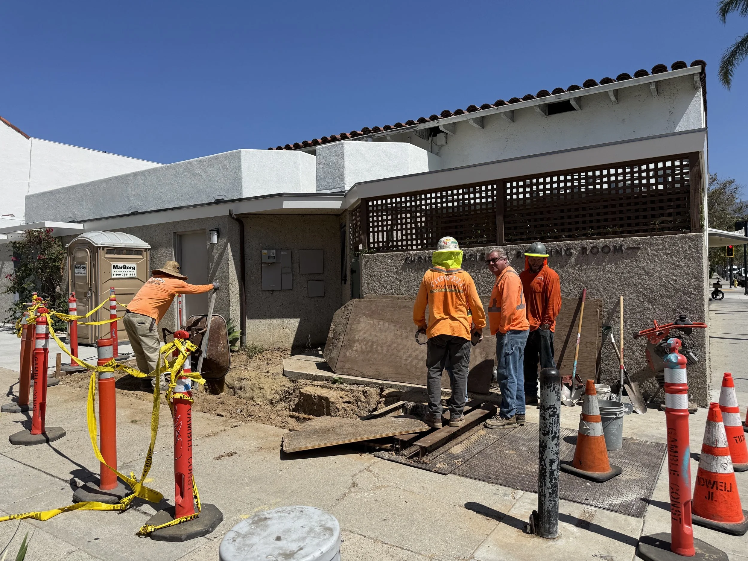  Wide shot of the Tidwell crew working outside the “…Science Reading Room” wall — open trench, traffic cones, and caution tape along the sidewalk. 