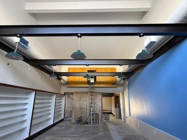  Wide view of the train room: green pendant lights hung from the black steel beams, white shelving along the left wall, and the yellow paneled loft at the far end. 