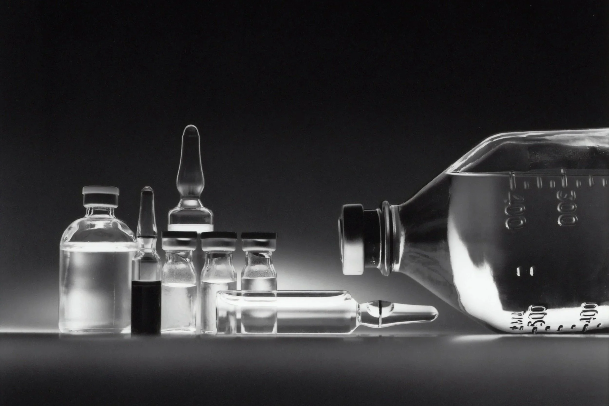 Black and white photograph of several glass medical vials and ampoules, with a large syringe lying on its side on a flat surface.