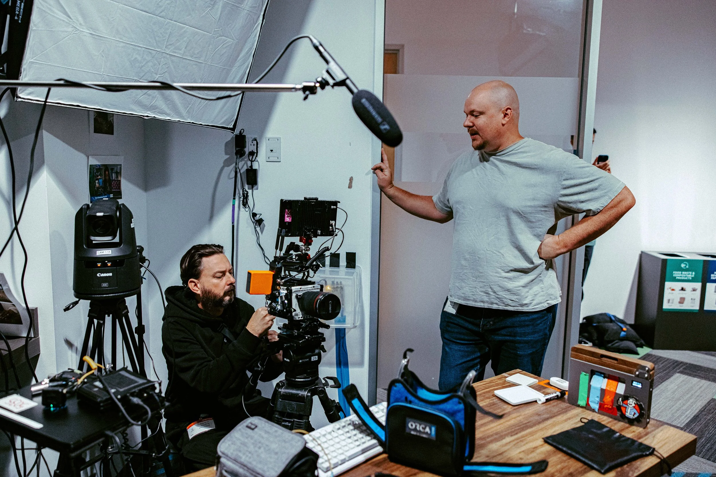 A man in a gray t-shirt standing and talking to a camera operator in a black hoodie, setting up a camera on a tripod in a room with filming equipment.