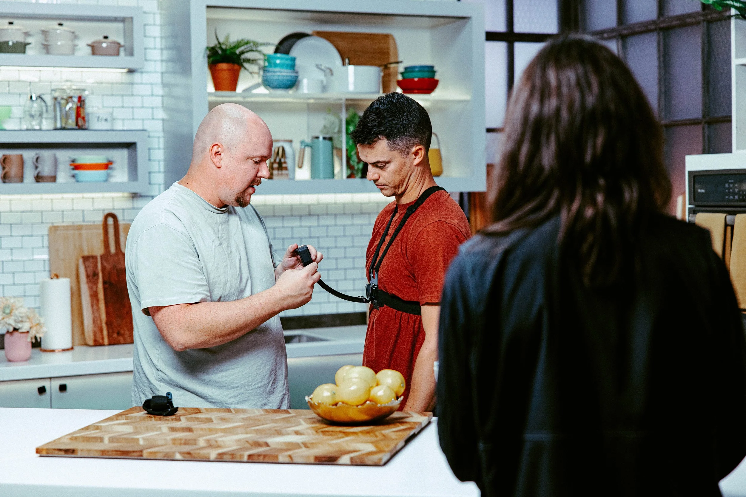 Three people in a kitchen, with two men looking at a device and a woman observing, in a modern kitchen with open shelves and various dishes in the background.