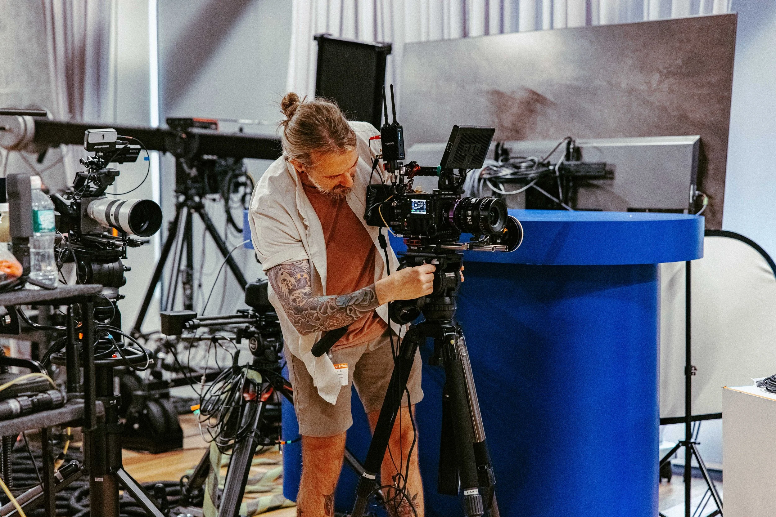 A man with tattoos on his arms is operating a professional video camera on a tripod in a studio with various equipment and cables around.