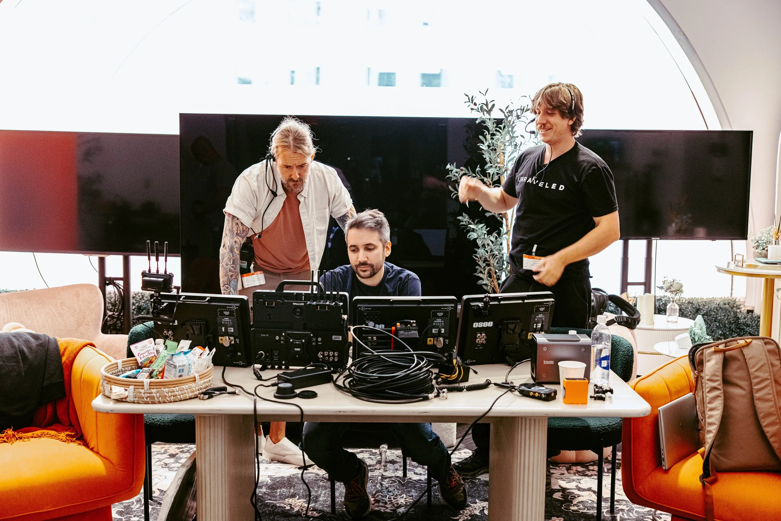 Three men working on video equipment in a modern office space with two large screens, a potted plant, and a colorful sofa.