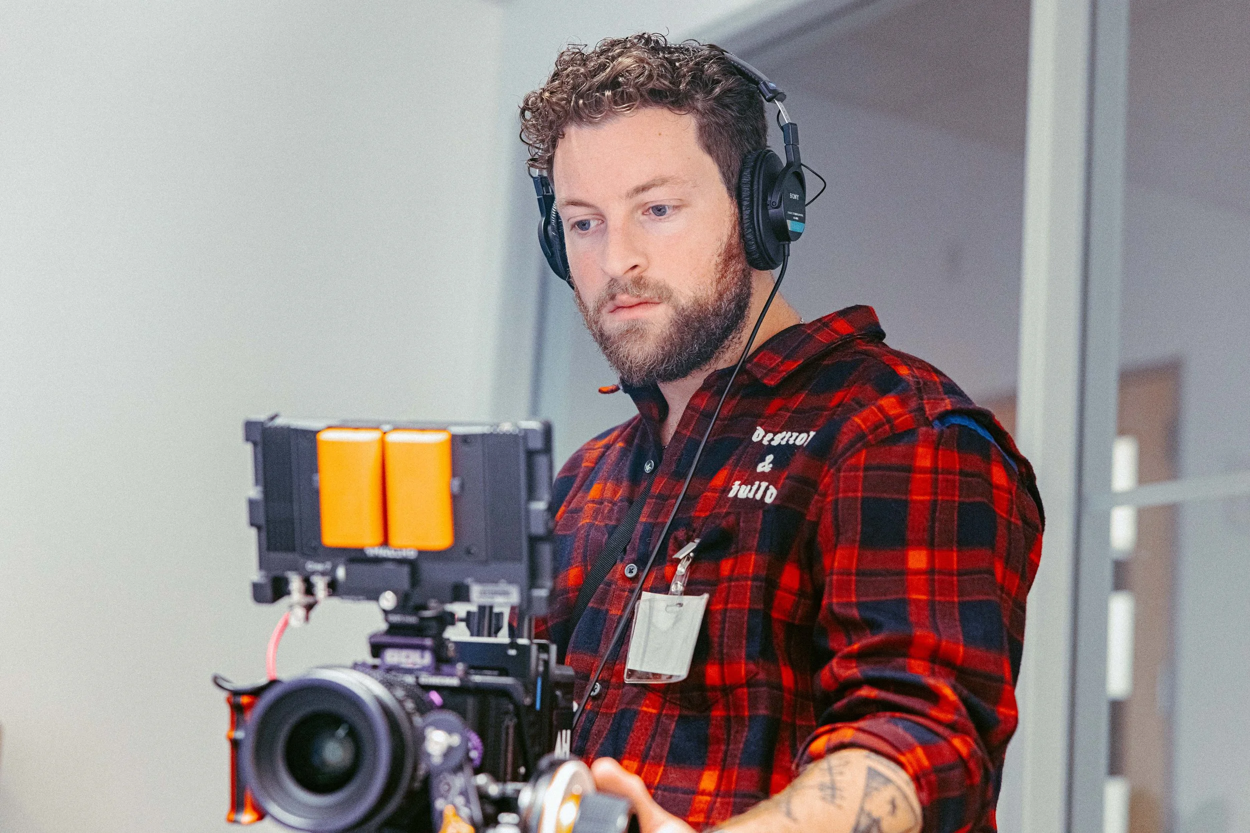 Man with curly hair and beard wearing headphones and a red plaid shirt operating a professional camera.