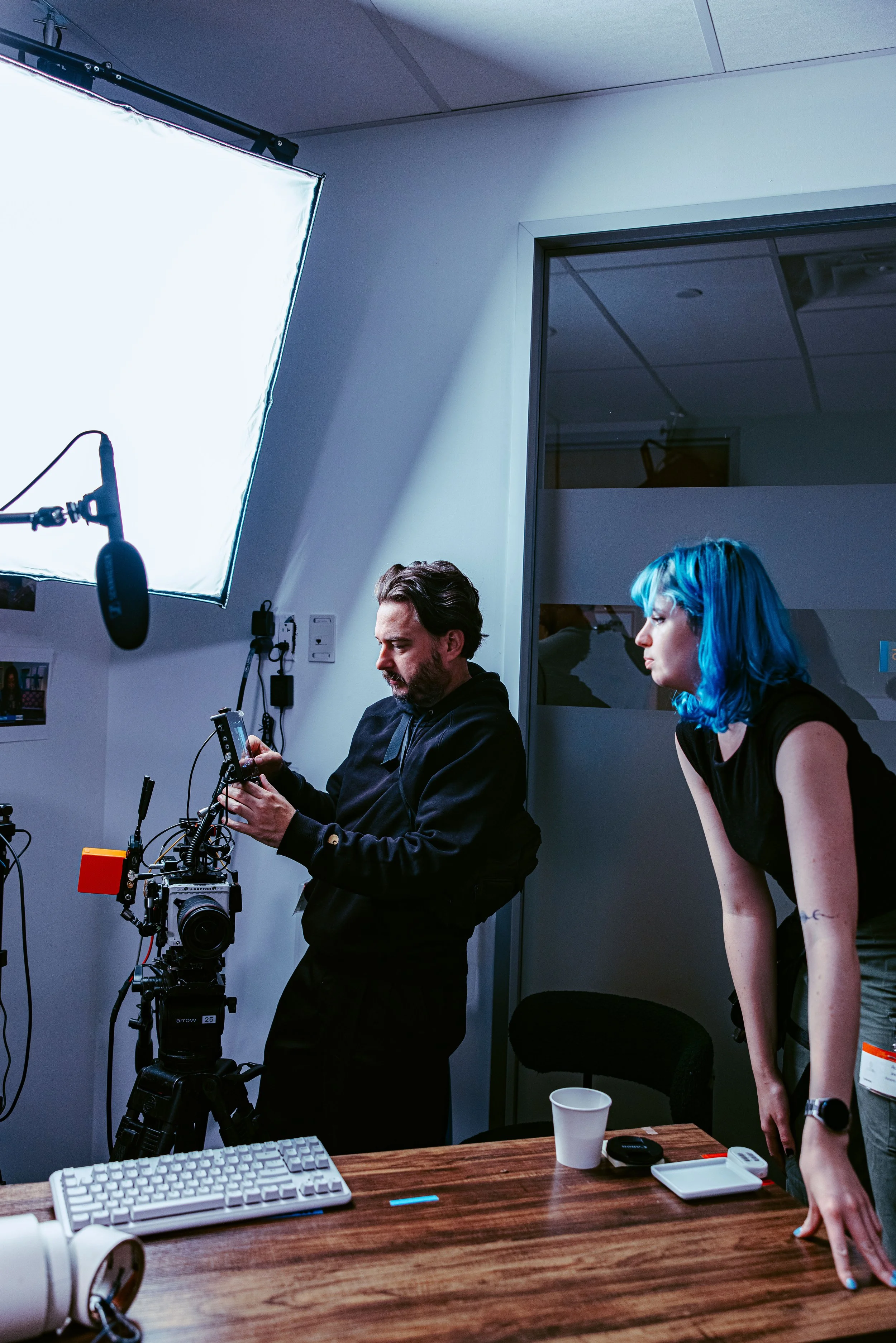 Two people in a behind-the-scenes setting with professional lighting and equipment. One person is looking at a phone, and the other is leaning on a table with a white cup and various items on it.