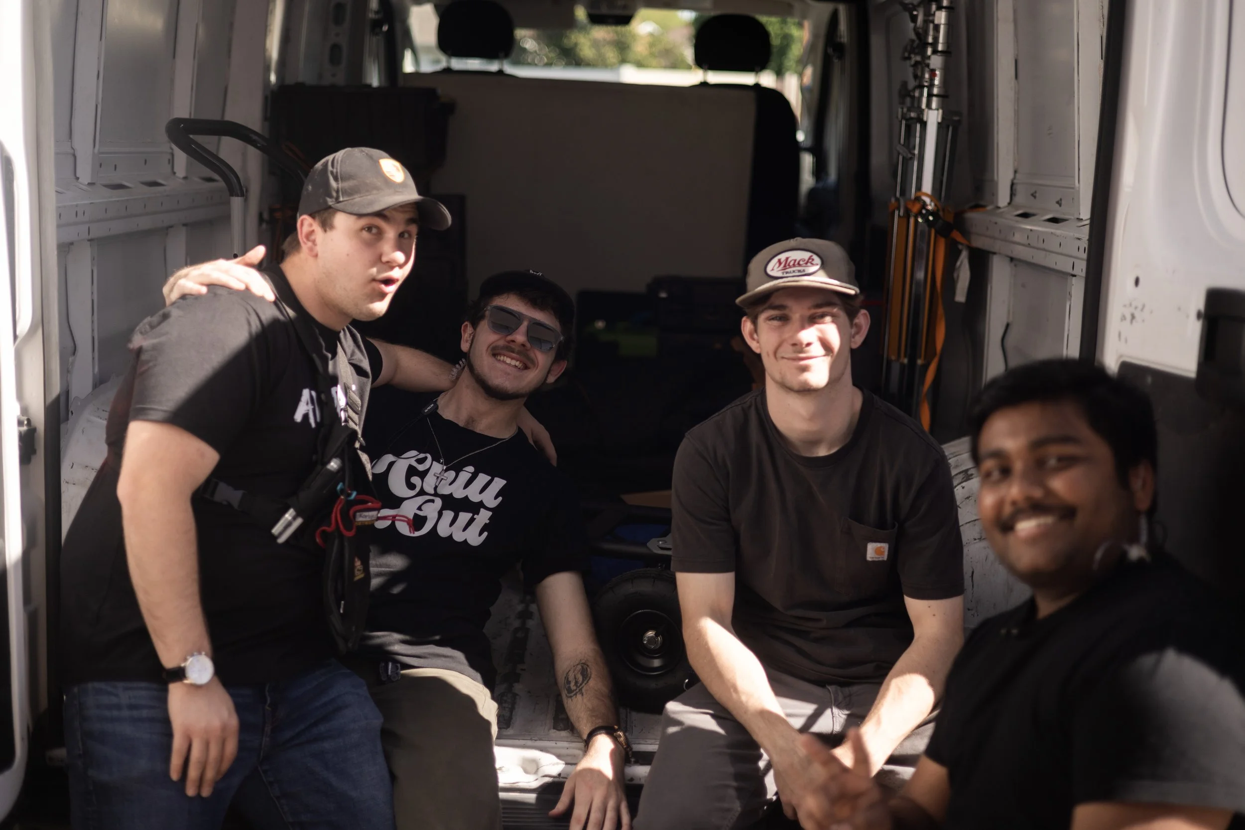 Four young men inside the back of a white van, smiling at the camera. The van is open, revealing sound equipment and other gear.