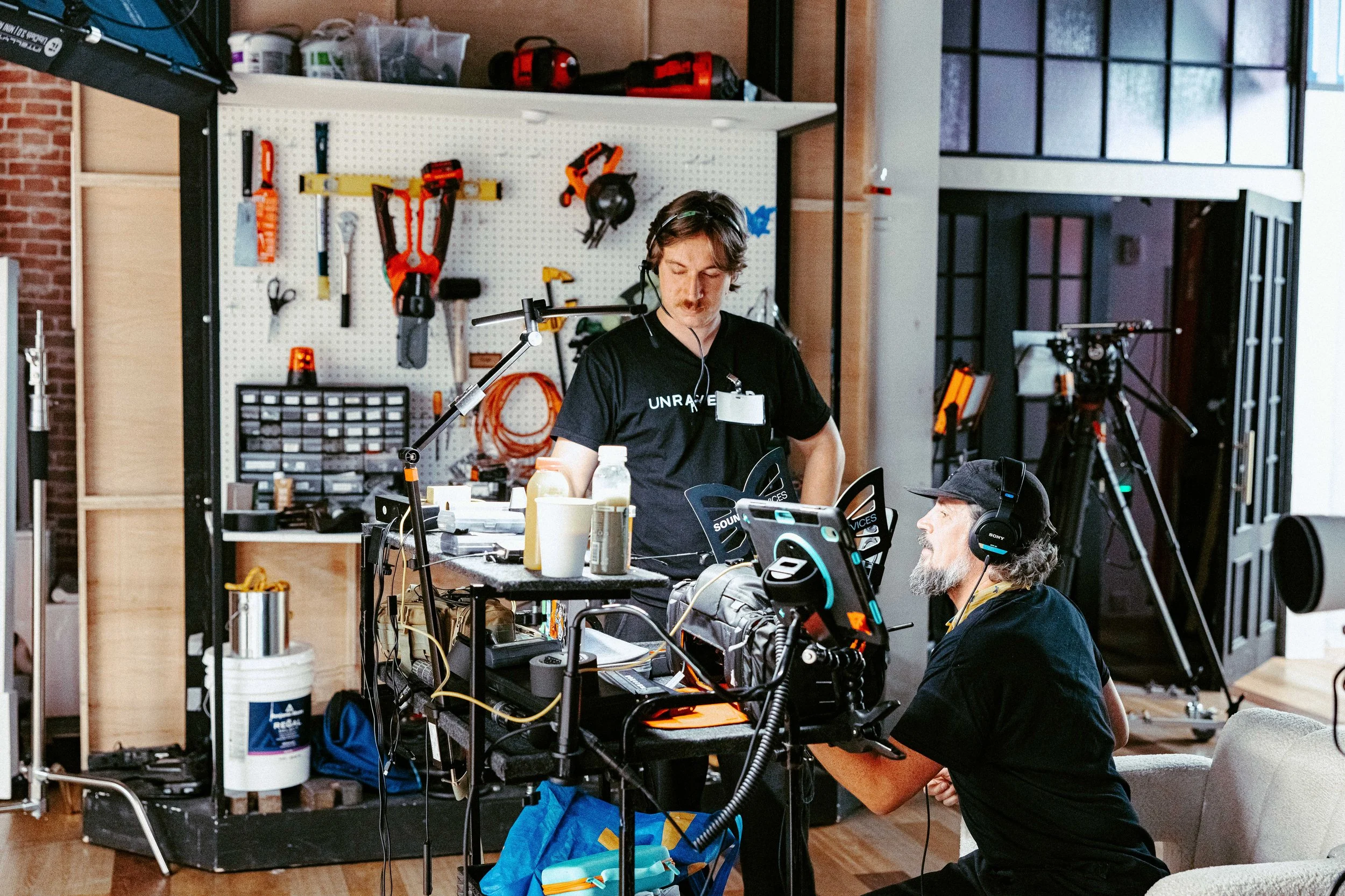 Two men working in a workshop with tools and equipment, one with a headset, the other with a cap, surrounded by cameras and technical gear.
