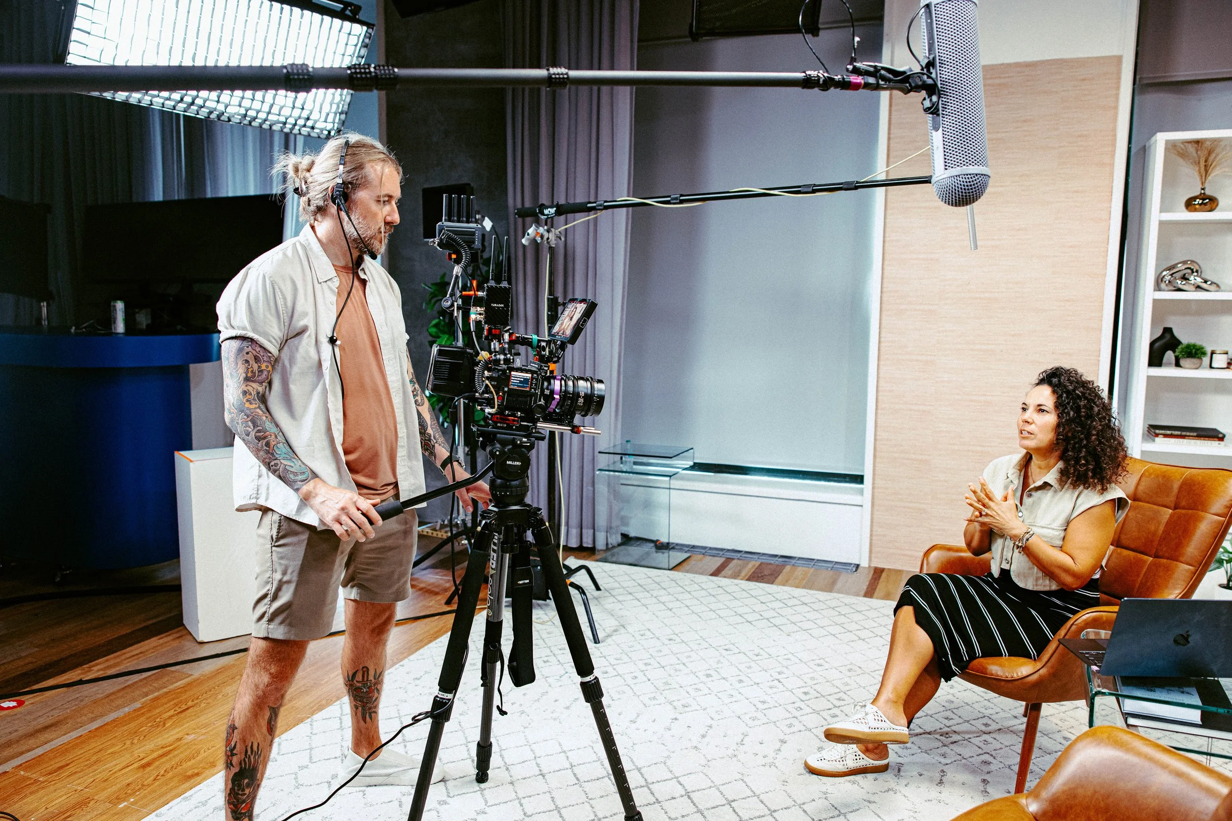 A man operating a professional video camera on a tripod records a woman sitting on a brown leather chair during an interview in a modern indoor setting with wooden floors, a white rug, gray curtains, and a bookshelf in the background.
