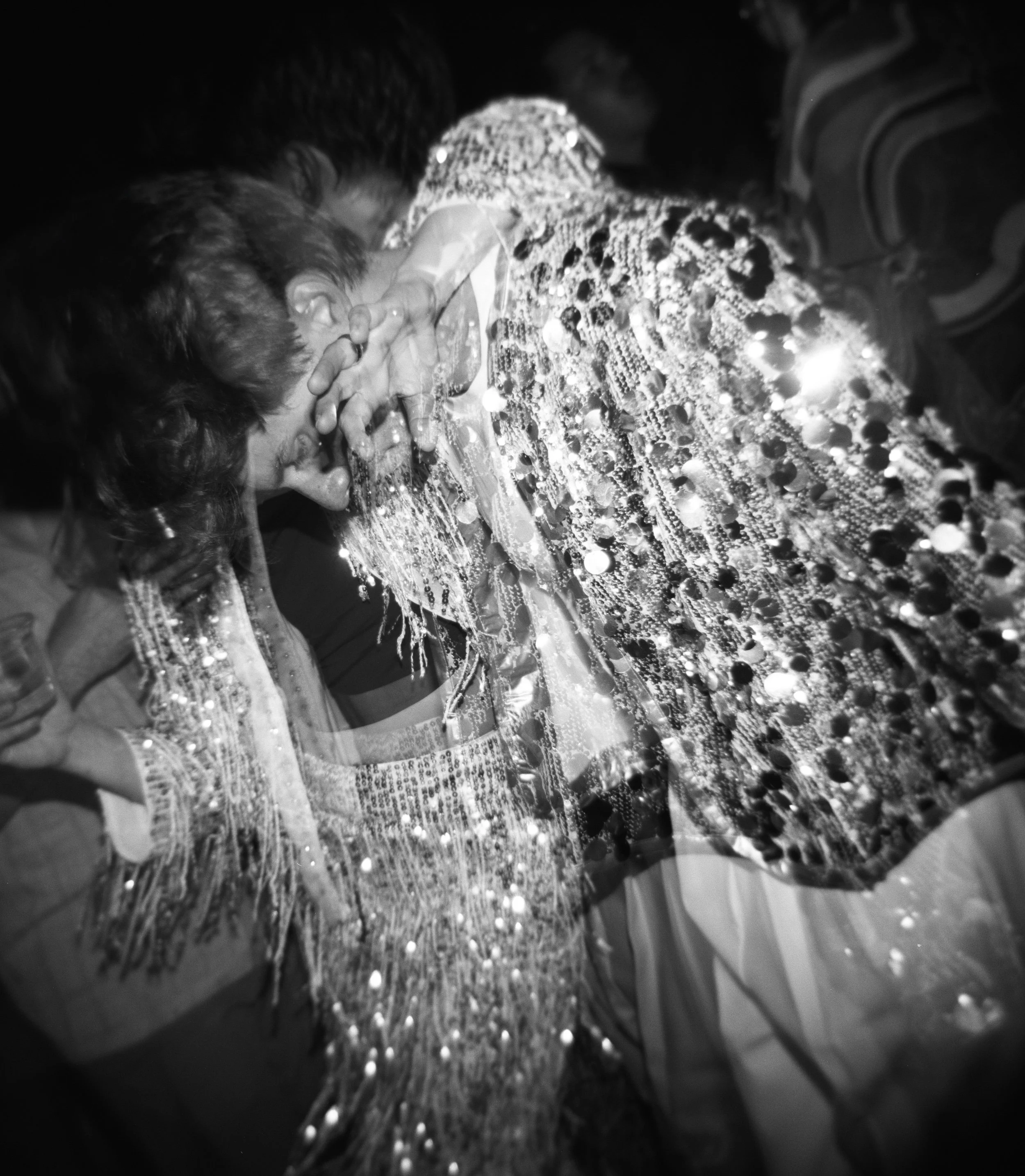A person in a highly decorated, shiny outfit, leaning in, seemingly crying or overwhelmed, with their hands covering their face, embraced by another person in a dark setting.